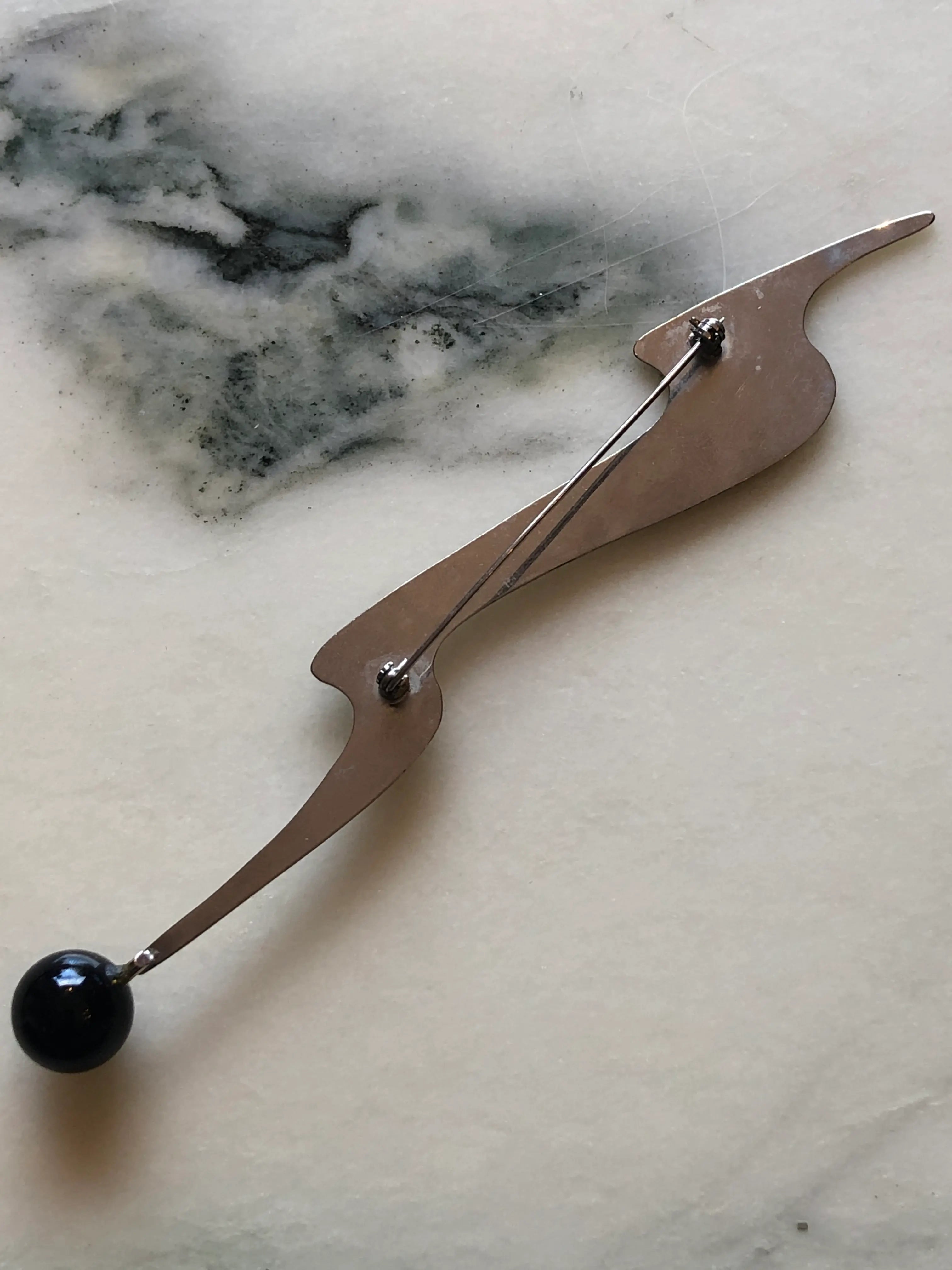 The back view of Broochella Midnight Comet vintage modernist brooch in polished silver-tone metal with an elongated abstract shape and glossy black spherical drop, measuring approximately 16.5 × 3 cm. Photographed on white and grey marble.