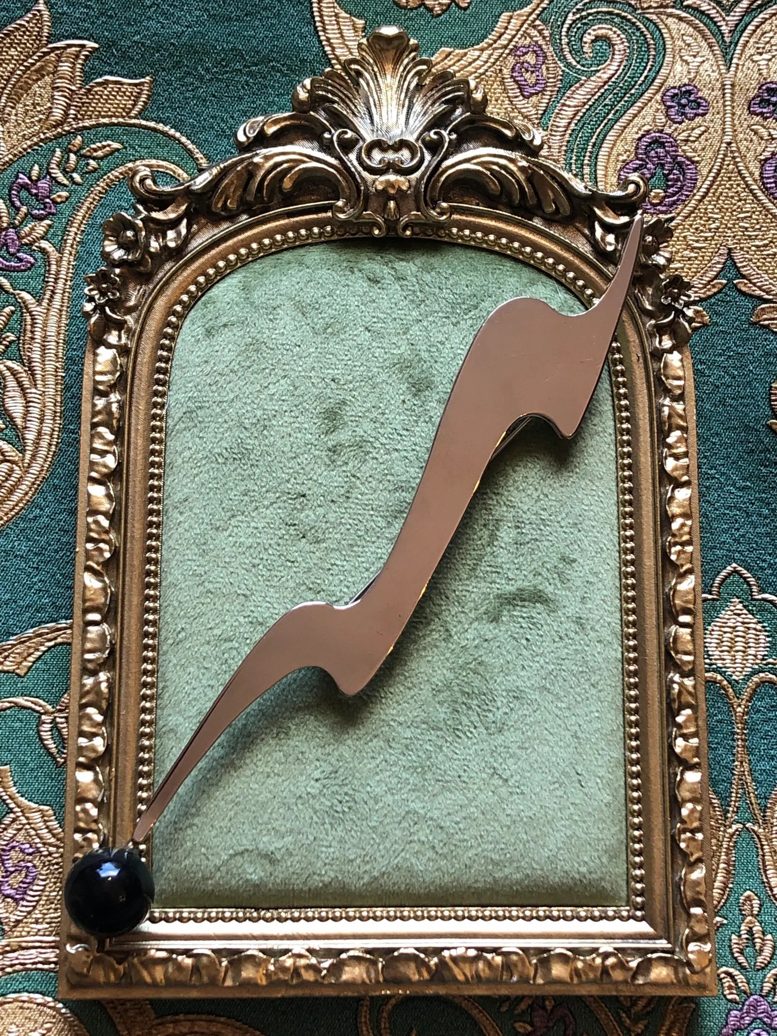 Broochella Midnight Comet vintage modernist brooch in polished silver-tone metal with an elongated abstract shape and glossy black spherical drop, measuring approximately 16.5 × 3 cm. Photographed on a green velvet tray with an ornate gold frame. The background is green and gold damask wallpaper