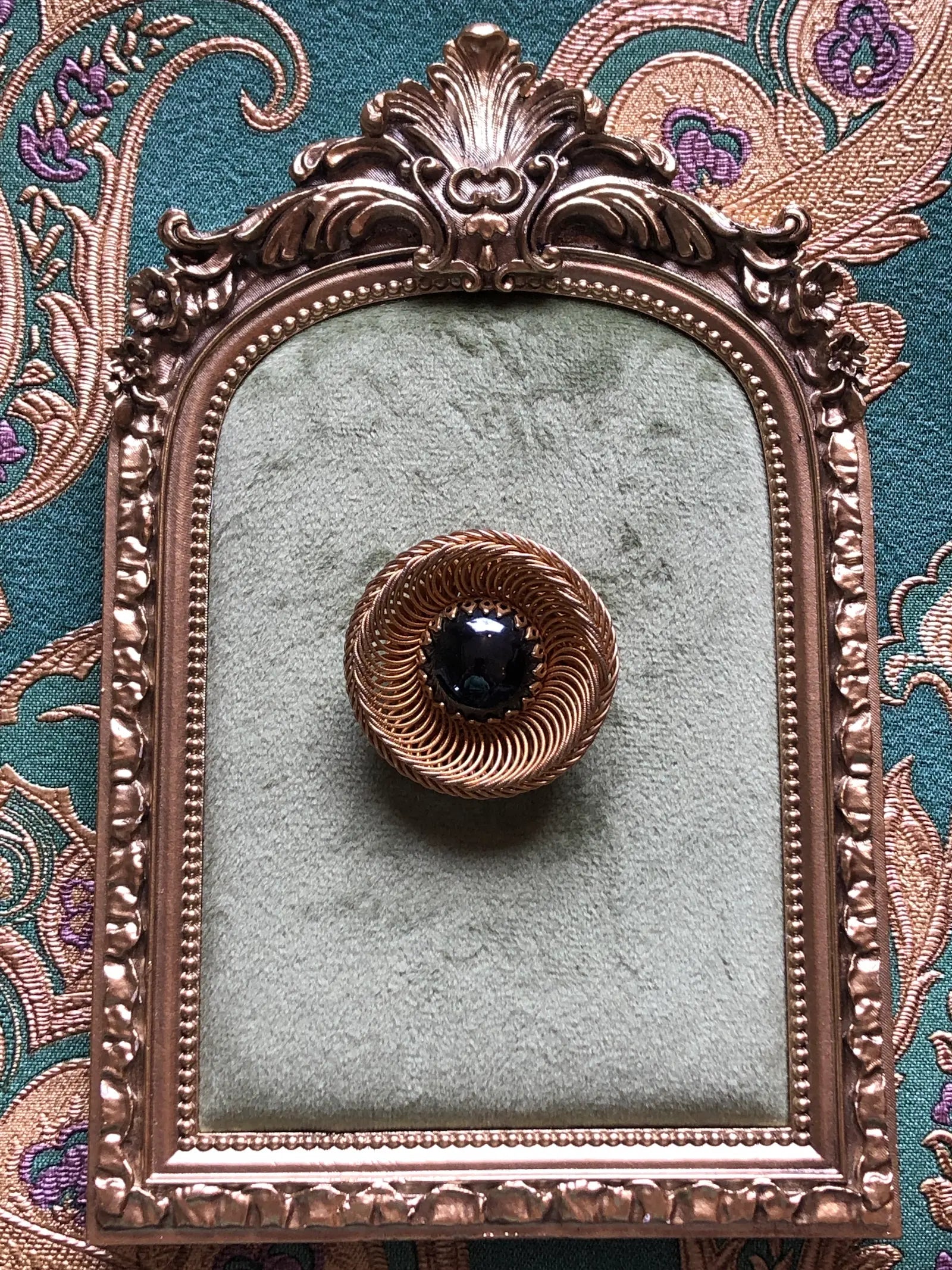 Broochella Midnight Halo vintage circular brooch with a glossy black domed cabochon and intricate spiral gold-tone wirework, measuring approximately 4 cm. Photographed on white a green velvet tray with an ornate gold frame. The background is green and gold damask wallpaper