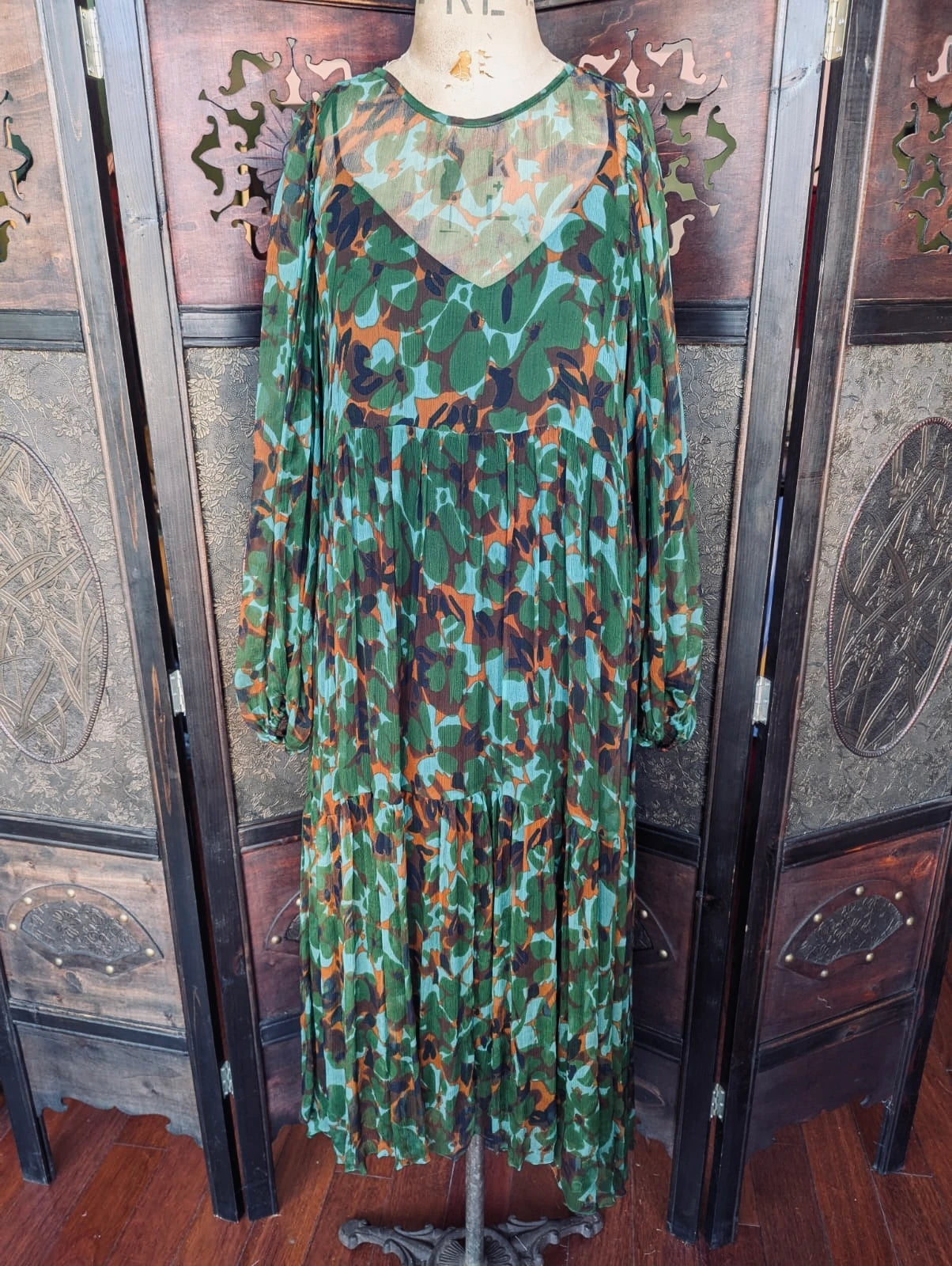 Broochella MLM Green,  blue, rust and black abstract floral, sheer, tiered, midi Dress with Slip – Size Small. Pictured on a mannequin. The background is an ornate, dark wood room divider. 
