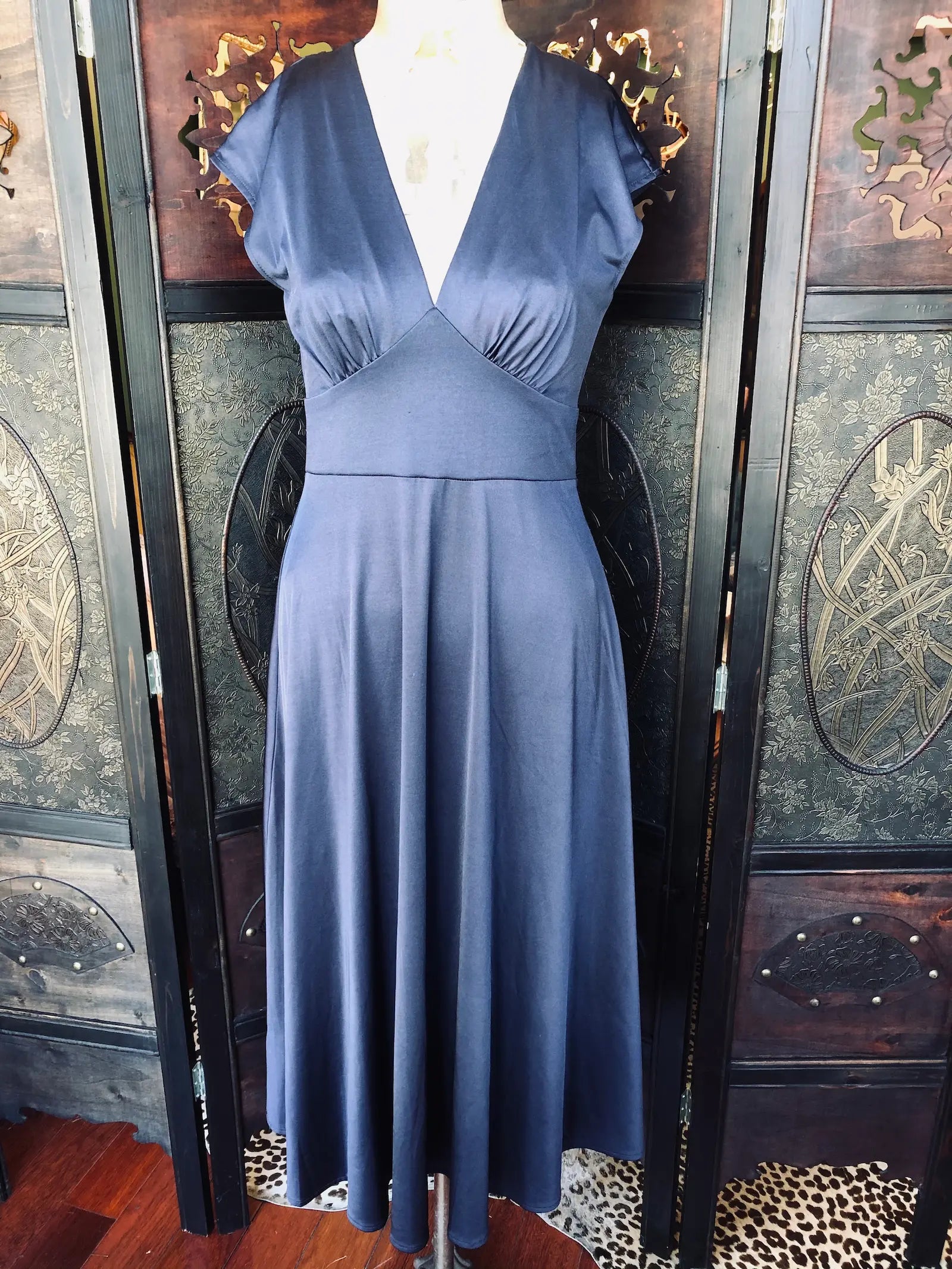 Broochella MNG size small, 92% Polyester, 8% Elastane, Navy
Deep V-neckline dress, with fitted bodice and  flowing skirt. It is midi length. It is presented on a vintage mannequin. The background is an ornate, dark wood room divider. 