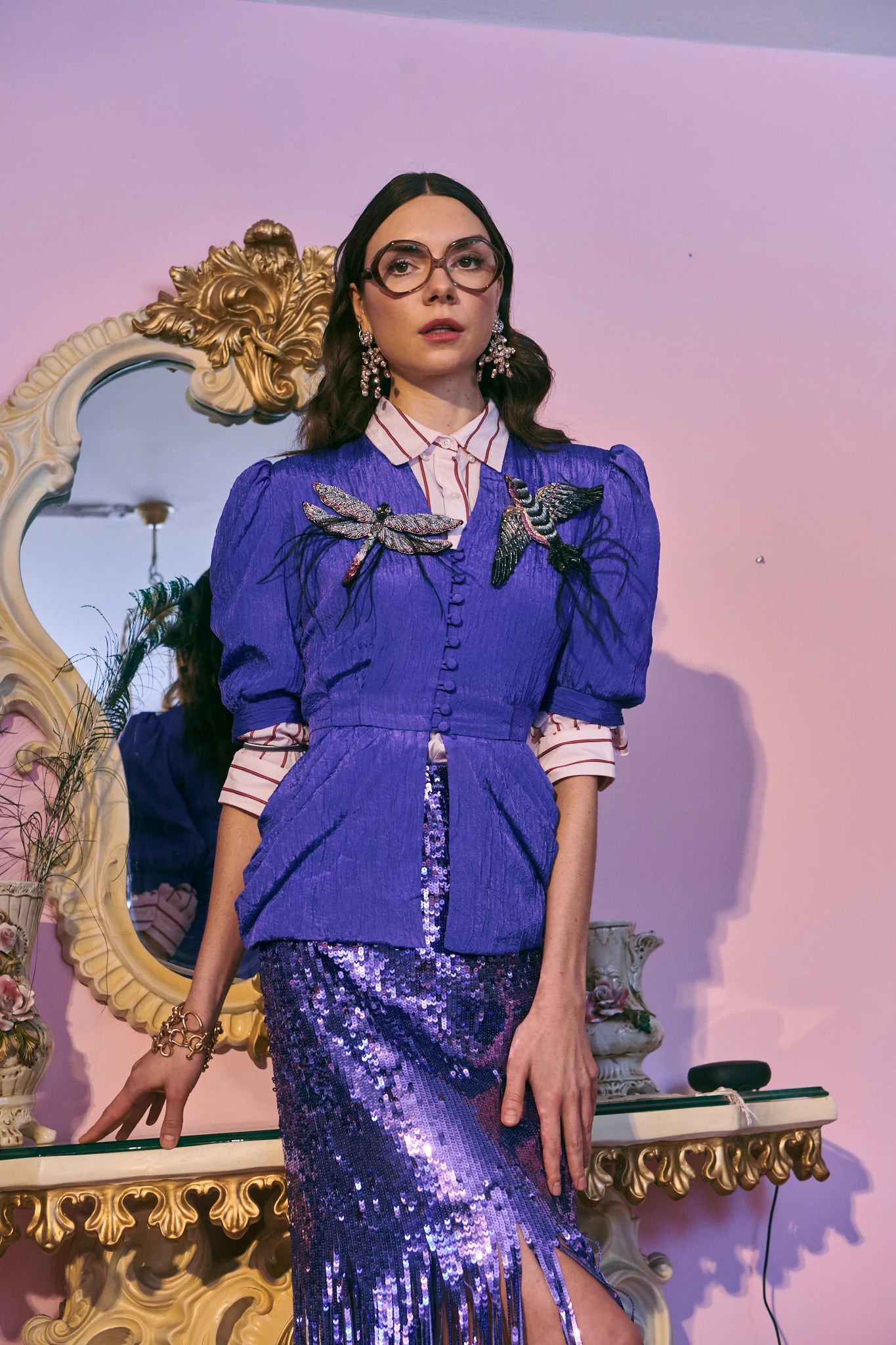 A model wearing a vibrant purple two-piece outfit consisting of a button-up jacket with the Broochellla Libellula dragonfly brooch and the Rondine hand beaded bird brooch.  She is wearing a  matching sequinned fringe skirt, posed in front of a pink wall and an ornate gold mirror.