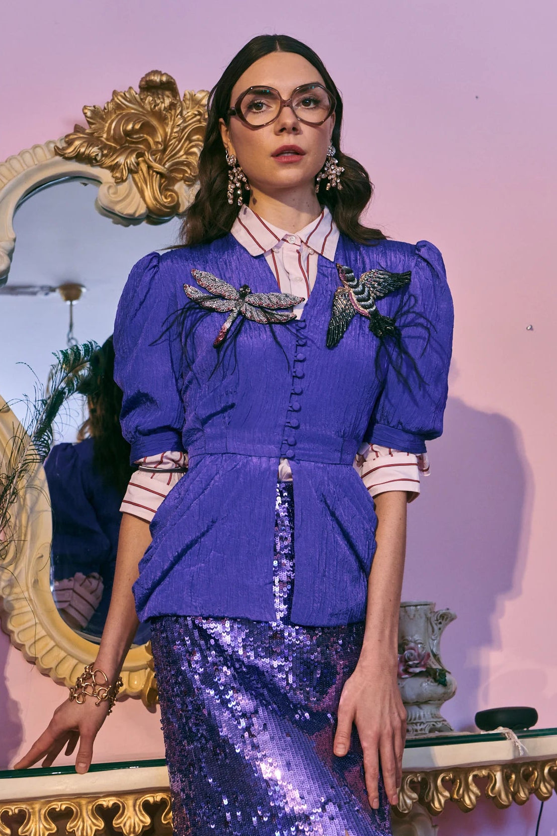 A model wearing a vibrant purple two-piece outfit consisting of a button-up jacket with the Broochella libellula  dragonfly brooch and the Broochella hand beaded Rondine bird brooch. She is wearing a matching sequinned fringe skirt,  and is posed in front of a pink wall and an ornate gold mirror.