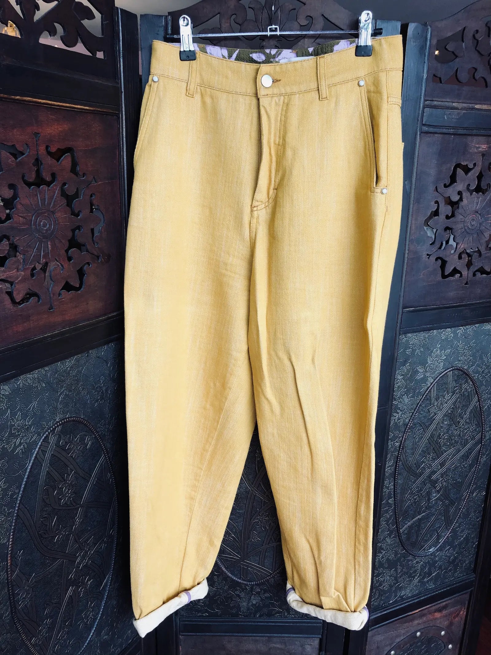 Broochella, The Stylist's Closest Momoni loose-fitting mustard yellow jeans. They are hanging from a silver hanger against a dark carved wooden screen.