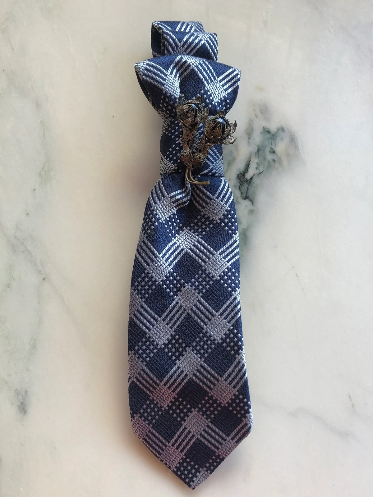 Broochella Montecristo Cravatta Tie Brooch Pin #10, a handmade designer statement brooch crafted from an upcycled vintage navy and silver geometric patterned tie, gathered and embellished with an ornate antique-style metal floral brooch, photographed on white marble.