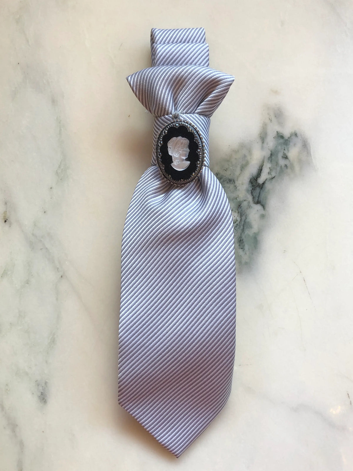 Broochella Montecristo Cravatta Tie Brooch Pin #17, a handmade designer statement brooch crafted from an upcycled vintage silver-grey striped tie, gathered and embellished with an oval black and white cameo framed in sparkling crystals, photographed on white marble.
