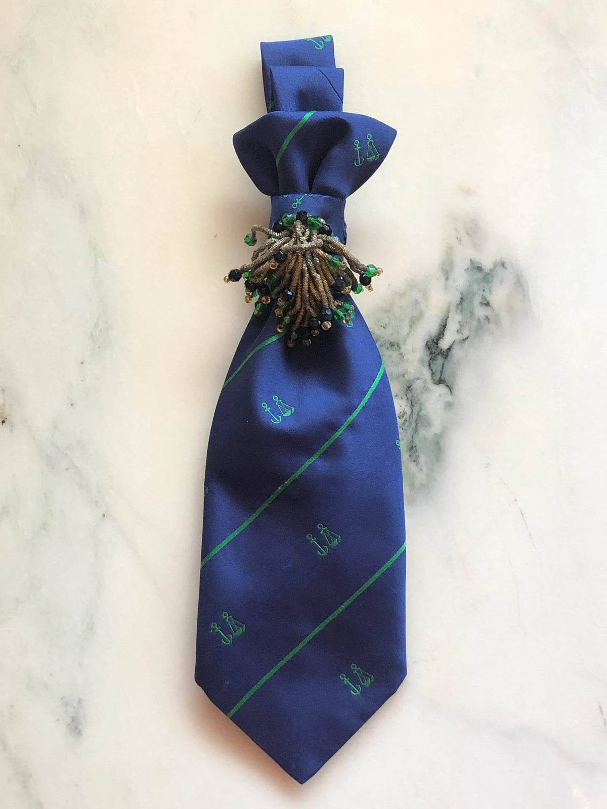 Broochella Montecristo Cravatta Tie Brooch Pin #19, a handmade designer statement brooch crafted from an upcycled vintage navy blue silk tie with green diagonal stripes and embroidered anchor motifs, finished with an ornate beaded embellishment in bronze, black and green, photographed on white marble.