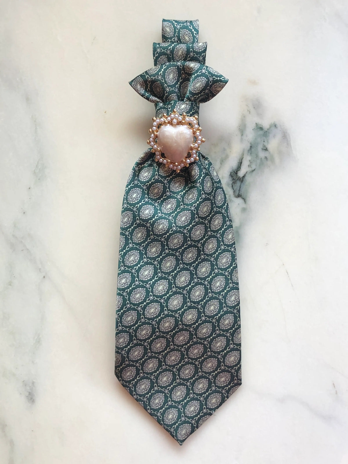 Broochella Montecristo Cravatta Tie Brooch Pin #20, a handmade designer statement brooch crafted from an upcycled vintage deep green patterned silk tie, finished with an ornate pearl heart embellishment in a gold-tone setting, photographed on white marble.