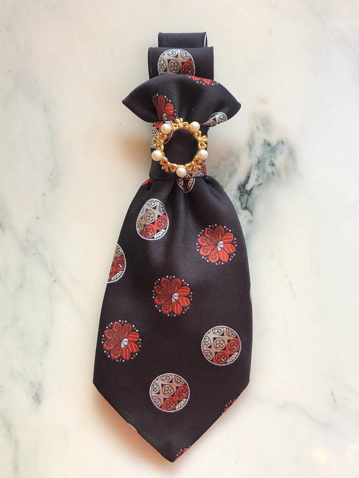 Broochella Montecristo Cravatta Tie Brooch Pin #23, a handmade designer statement brooch created from an upcycled vintage brown silk tie with red and ivory woven motifs, finished with a gold-tone circular embellishment and pearl details, photographed on white marble.