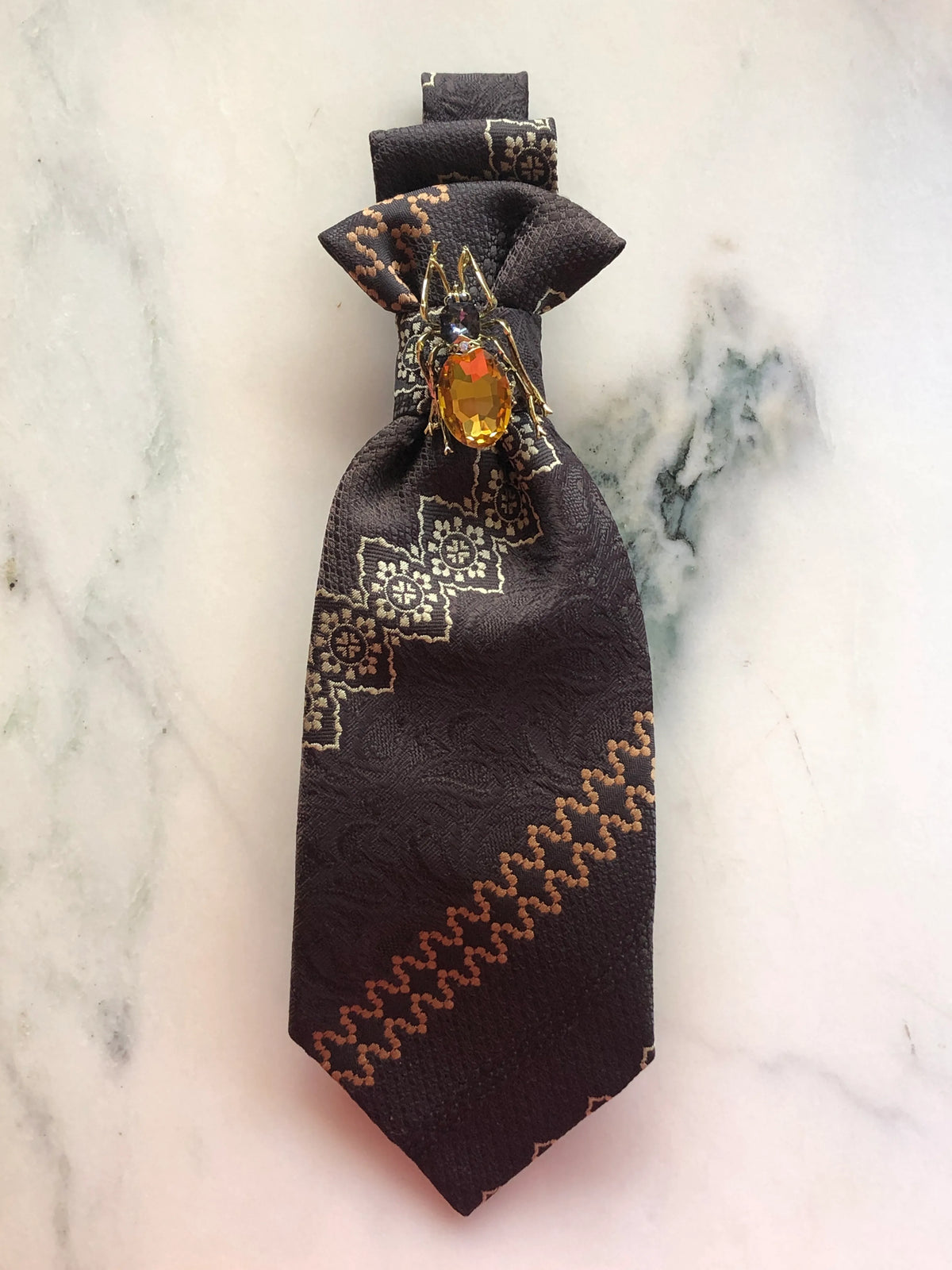 Broochella Montecristo Cravatta Tie Brooch Pin #25, a handmade designer statement brooch crafted from an upcycled vintage dark brown patterned tie, embellished with a gold-tone insect jewel featuring a faceted amber crystal, photographed on white marble.
