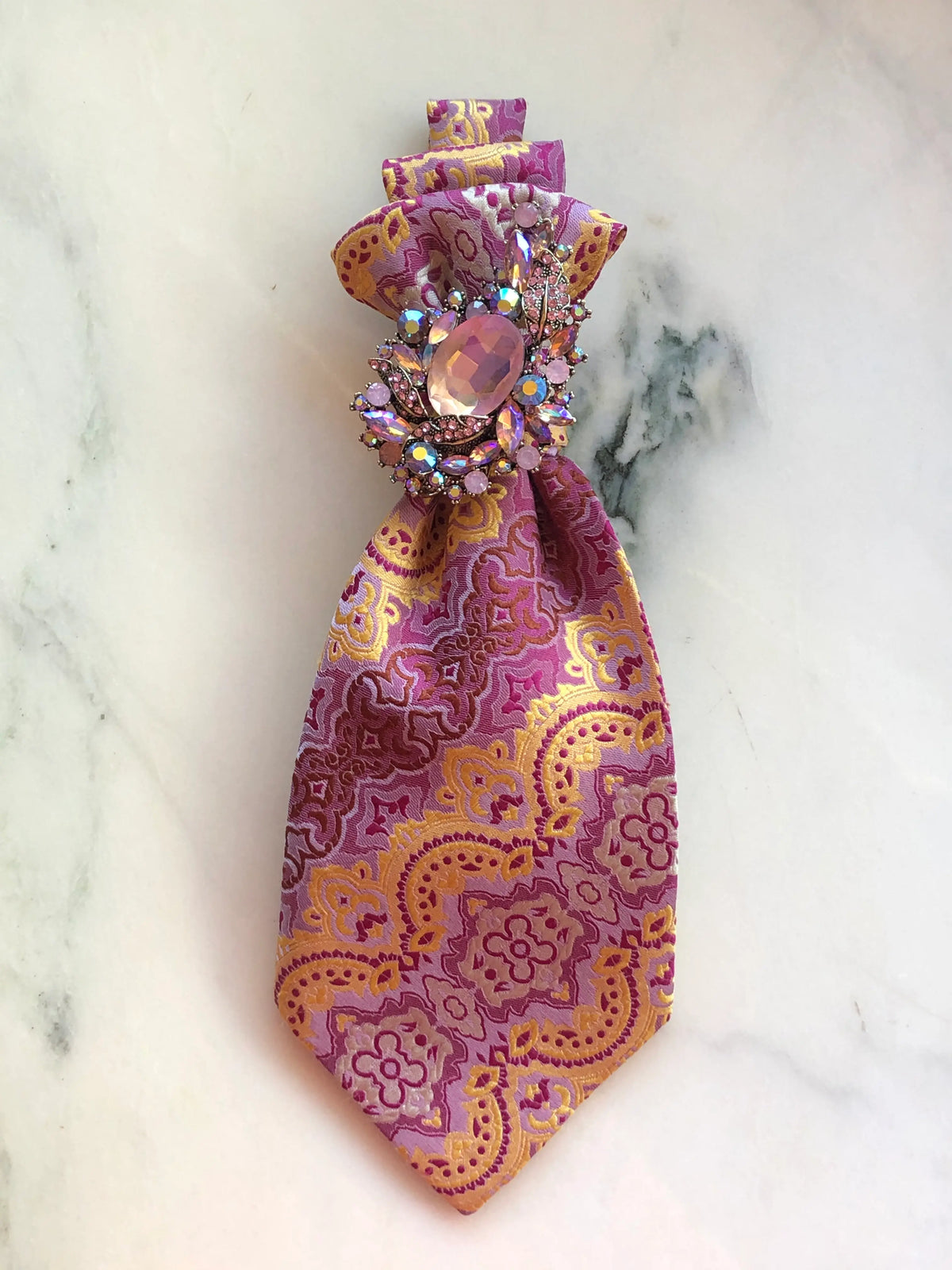 Broochella Montecristo Cravatta Tie Brooch #7, a handmade designer statement brooch created from an upcycled vintage pink, gold and mauve patterned tie, finished with an ornate pink crystal and rhinestone jewel, photographed on white marble.