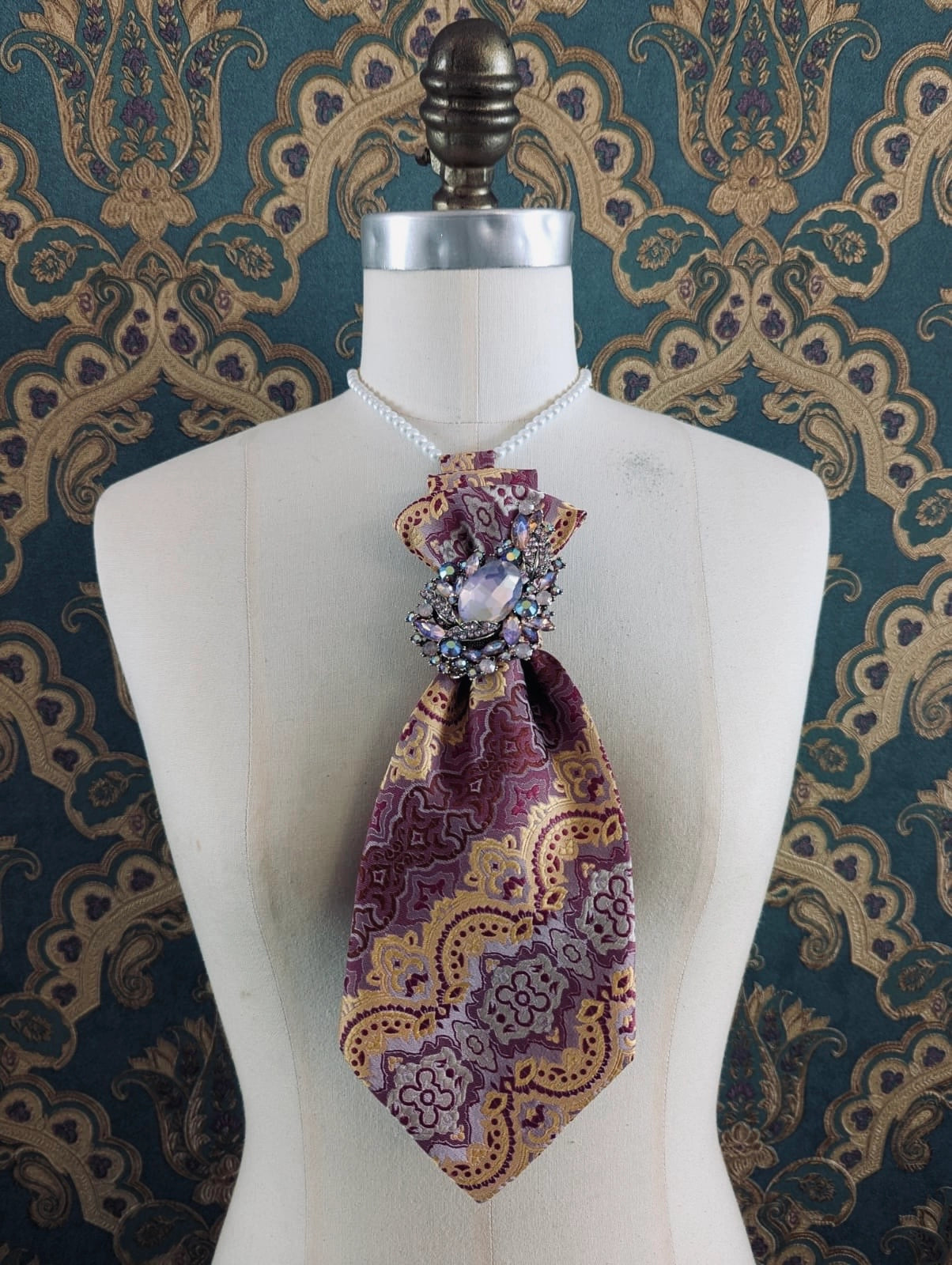 Broochella Montecristo Cravatta tie brooch. Made from up cycled vintage ties and embellished with a vintage brooch. Pictured on a mannequin. The background is green and gold damask wallpaper.