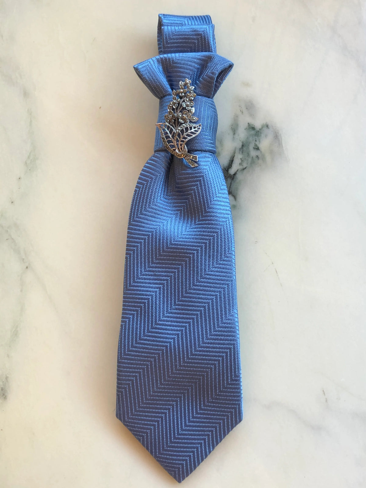 Broochella Montecristo Cravatta Tie Brooch Pin #13, a handmade designer statement brooch crafted from an upcycled vintage blue herringbone tie, embellished with an ornate silver floral brooch, photographed on white marble.