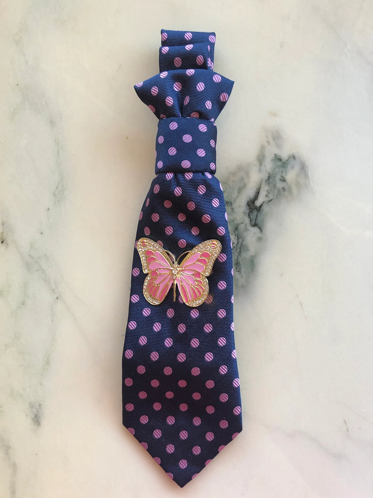 Broochella Montecristo Cravatta Tie Brooch Pin #14, a handmade designer statement brooch crafted from an upcycled vintage navy tie with pink polka dots, embellished with a pink and gold crystal butterfly brooch, photographed on white marble.