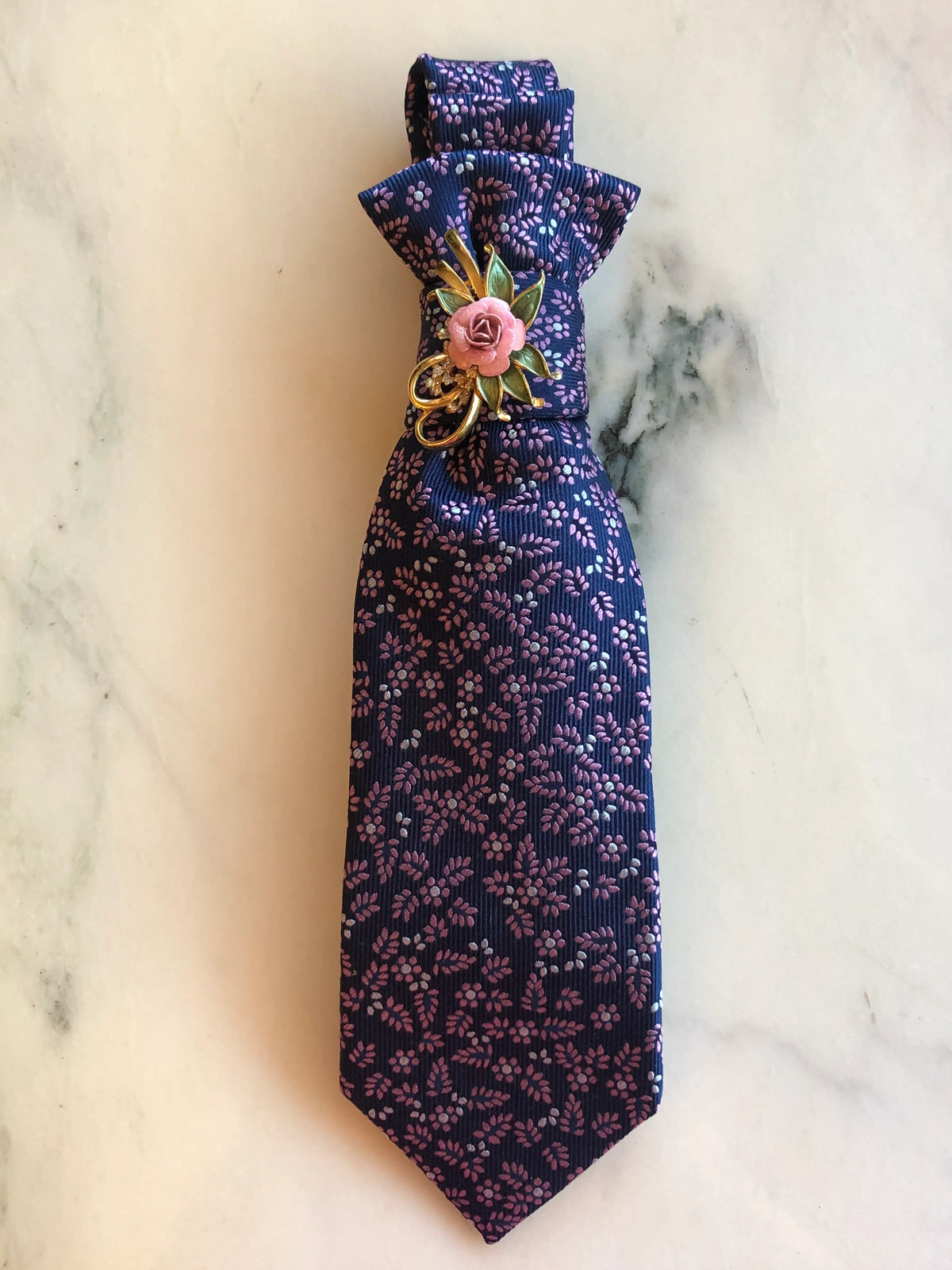 Broochella Montecristo Cravatta Tie Brooch Pin #15, a handmade designer statement brooch crafted from an upcycled vintage navy tie with delicate pink floral detailing, finished with a vintage-style pink rose and gold floral embellishment, photographed on white marble.