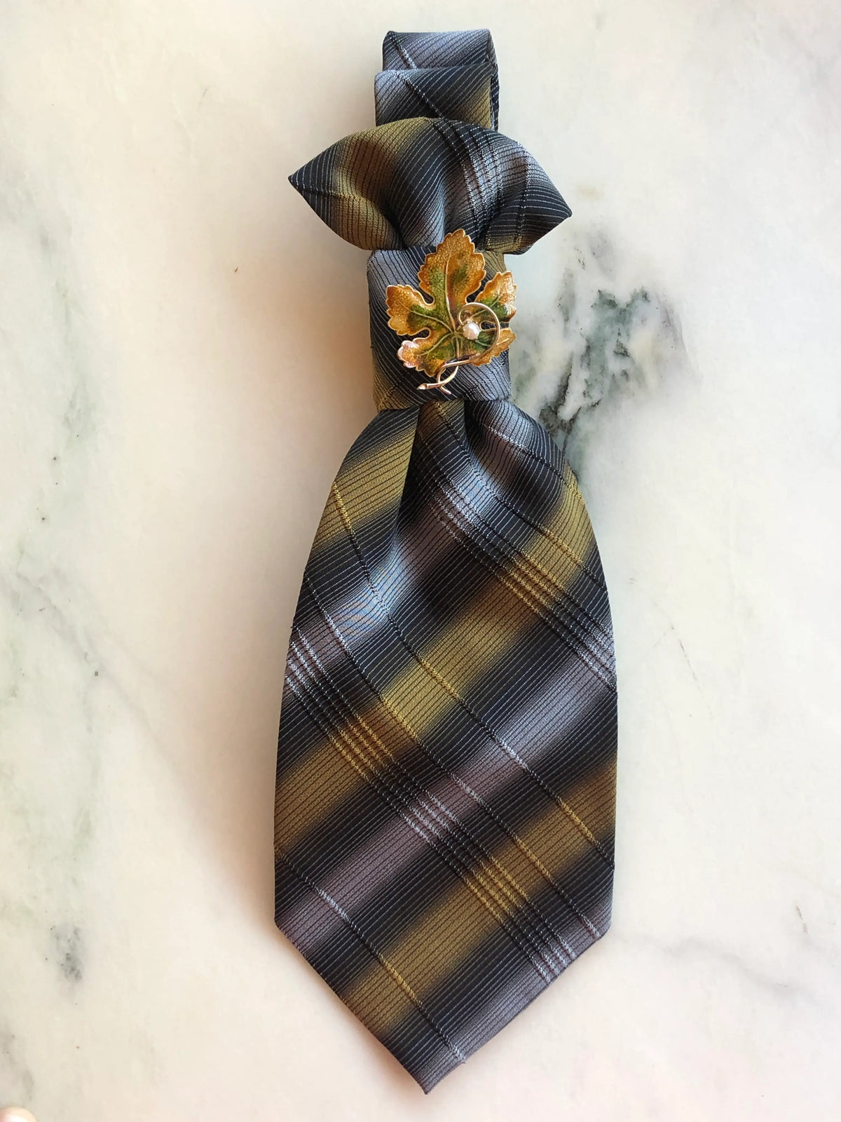 Broochella Montecristo Cravatta Tie Brooch Pin #4, a handmade designer statement brooch crafted from an upcycled vintage grey, charcoal and antique gold striped tie, finished with an ornate gold and green enamel leaf brooch with pearl detail, photographed on white marble.