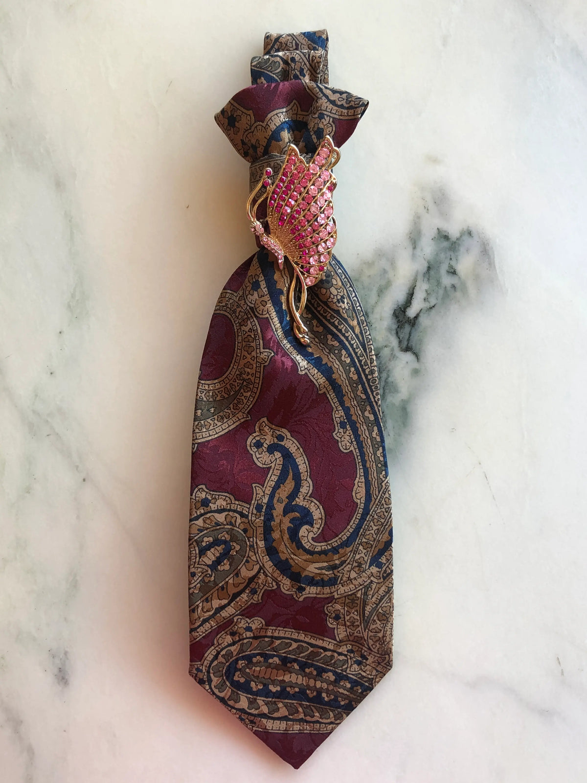 Broochella Montecristo Cravatta Tie Brooch Pin #5, a handmade designer statement brooch crafted from an upcycled vintage burgundy, navy and taupe paisley tie, embellished with a gold-tone wing brooch set with pink crystals, photographed on white marble.