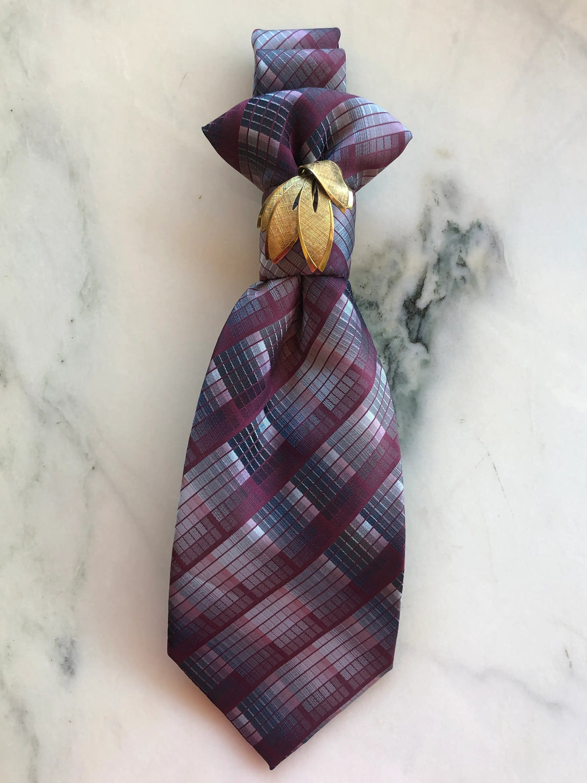 Broochella Montecristo Cravatta Tie Brooch Pin #6, a handmade designer statement brooch crafted from an upcycled vintage burgundy, silver-grey and navy plaid tie, finished with a sculptural gold-tone leaf brooch, photographed on white marble.