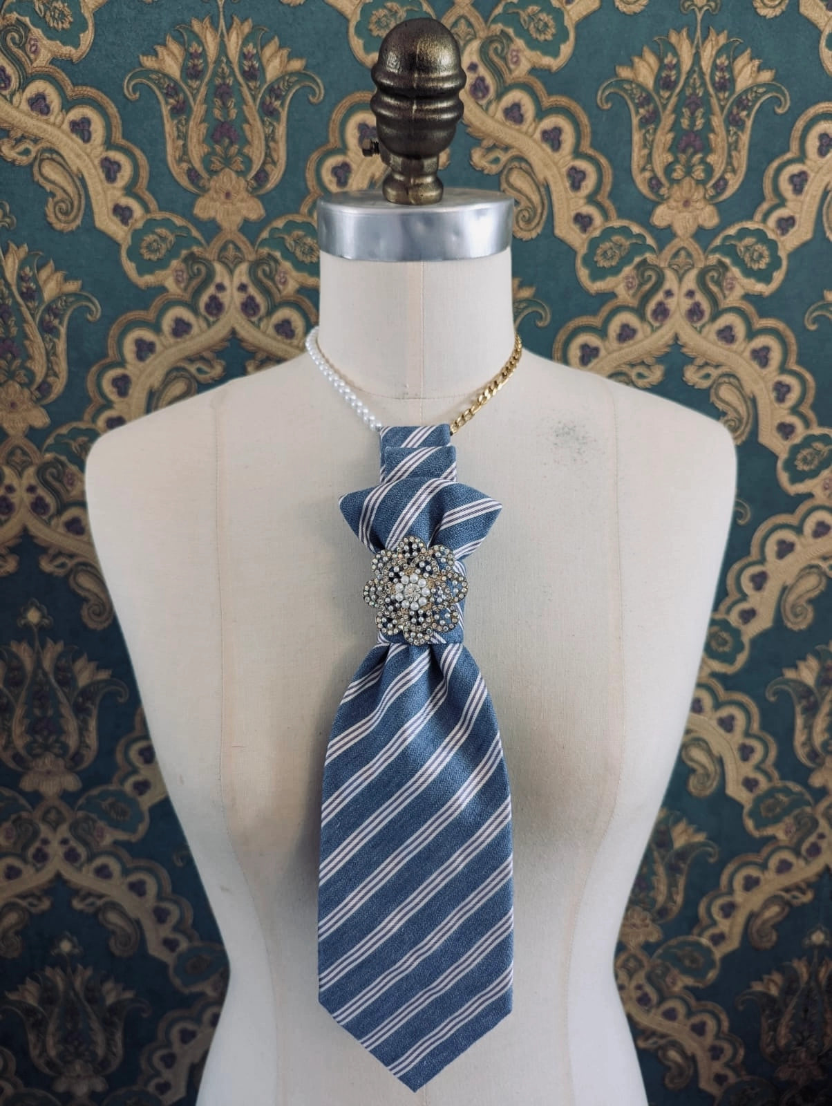 A Broochella Montecristo cravatta upcycled vintage blue and white striped tie brooch with a jeweled silver-toned knot, displayed on a cream-colored dress form mannequin against a dark teal and gold patterned wallpaper background.