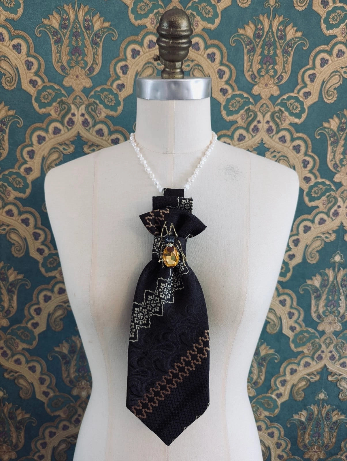 Broochella Montecristo cravatta 25 vintage up-cycled tie brooch in brown, orange and white 70s pattern. Pinned to the knot is a vintage brooch. It is pictured on a mannequin. The background is green and gold damask wallpaper. 