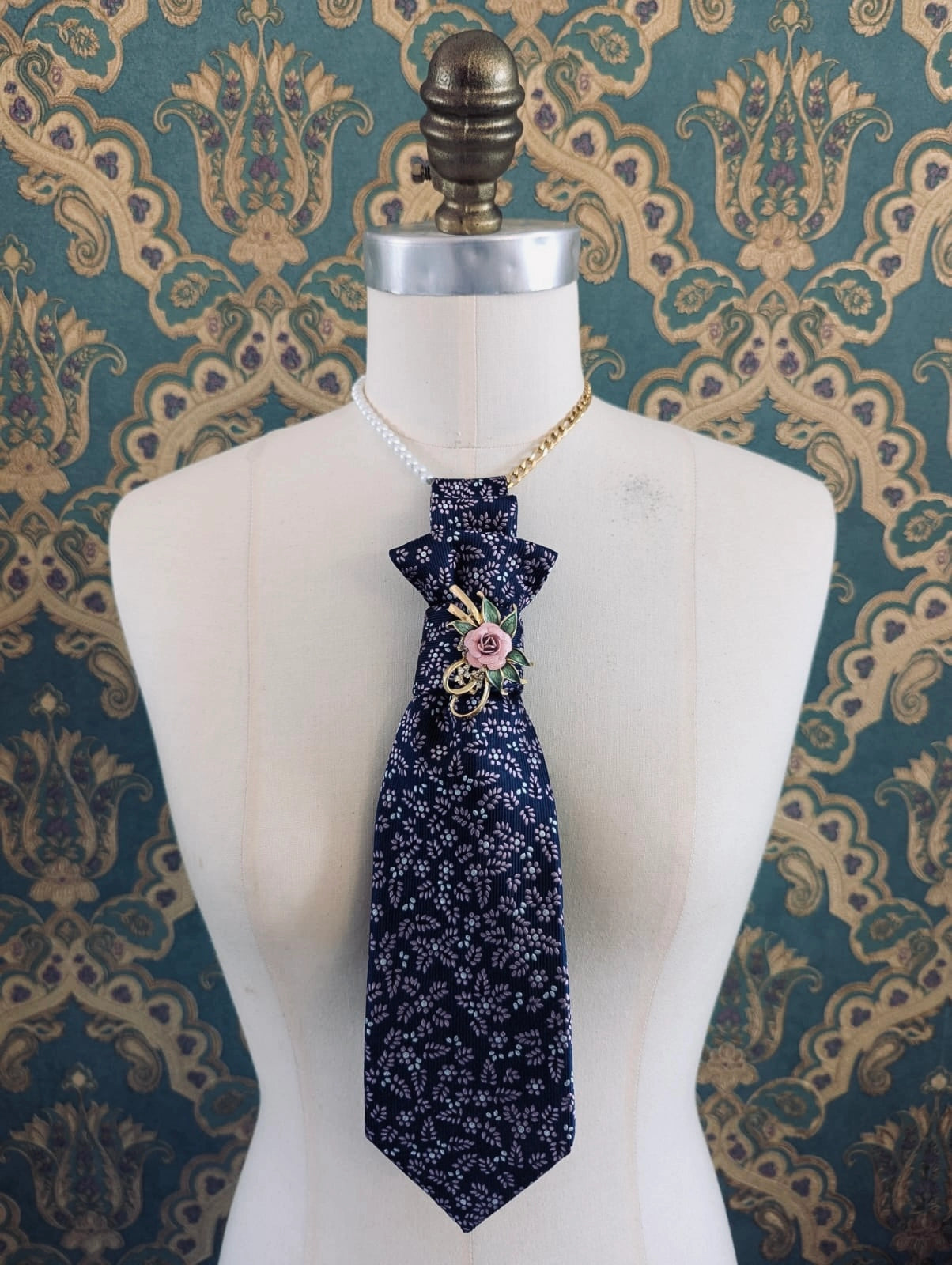 A Broochella montecristo navy blue patterned vintage up-cycled tie brooch, with a vintage brooch embellishment. It is displayed on a white dress form mannequin, styled with a gold chain and pearl necklace against a teal and gold damask wallpaper background.