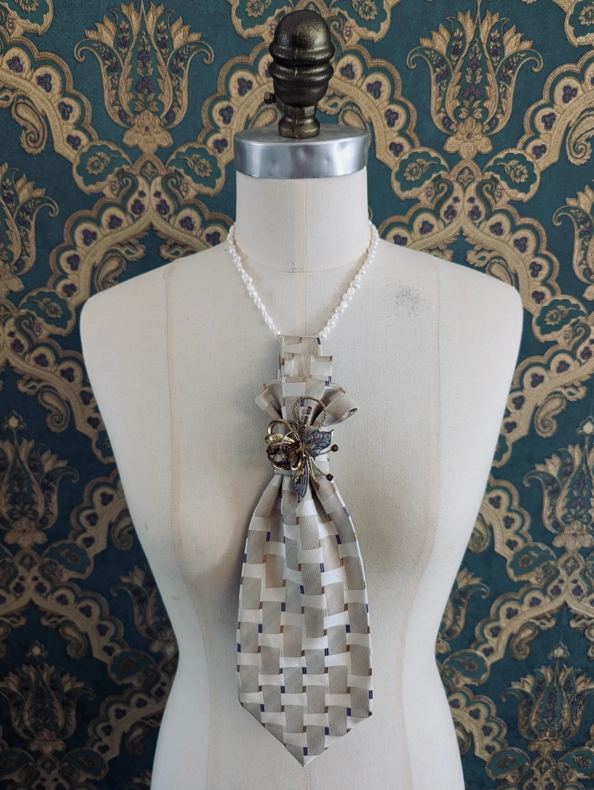 A Broochella Montecristo cravatta number 22 upcycled vintage tie brooch in bige herringbone weave pattern, with a vintage brooch embellishment. It is pinned on a cream-coloured dress form mannequin. The background is green and gold damask wallpaper. 