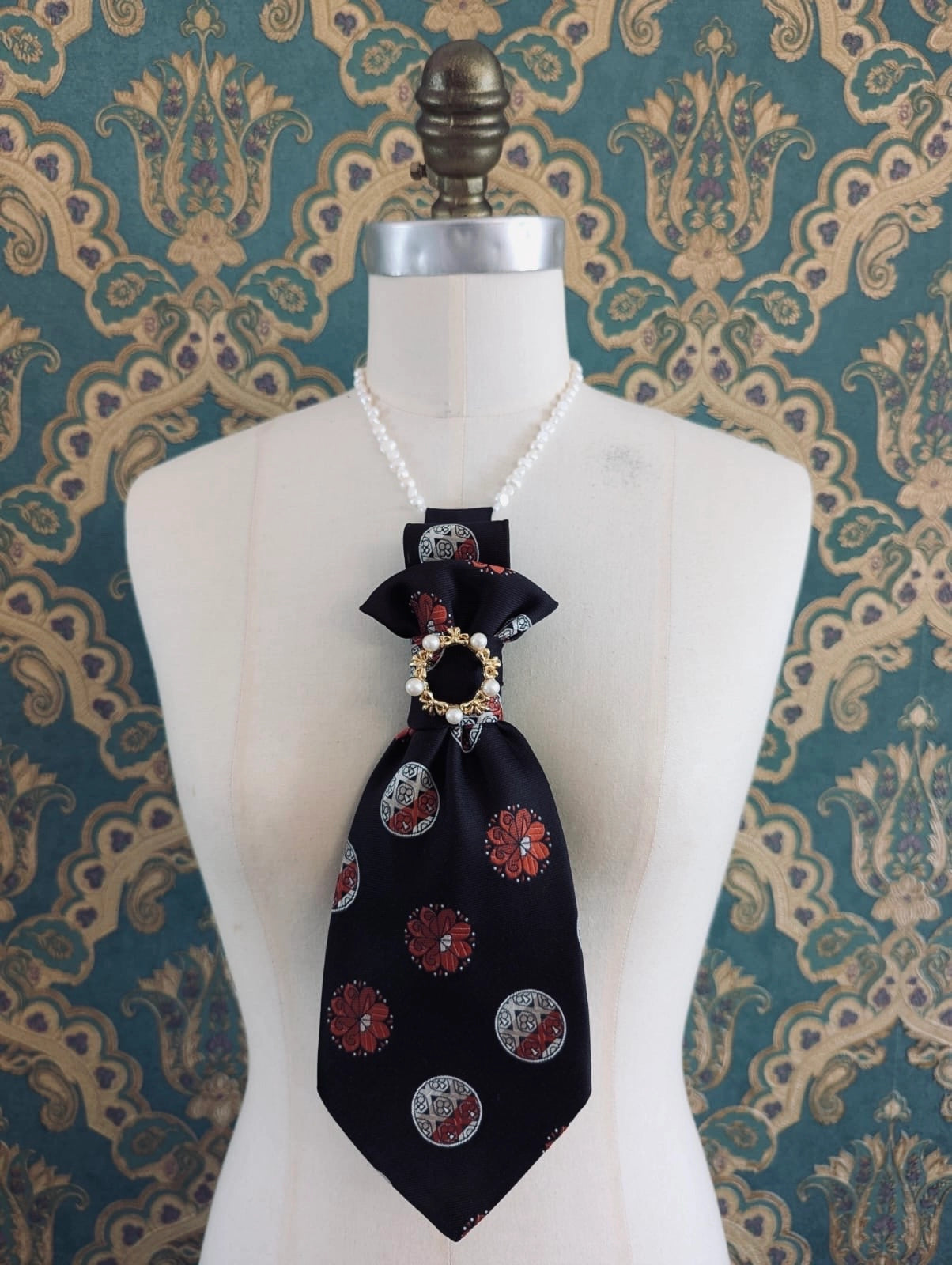 A Broochella Montecristo cravatta number 23 upcycled vintage tie brooch in black and red corcular pattern, with a vintage brooch embellishment. It is pinned on a cream-coloured dress form mannequin. The background is green and gold damask wallpaper. 