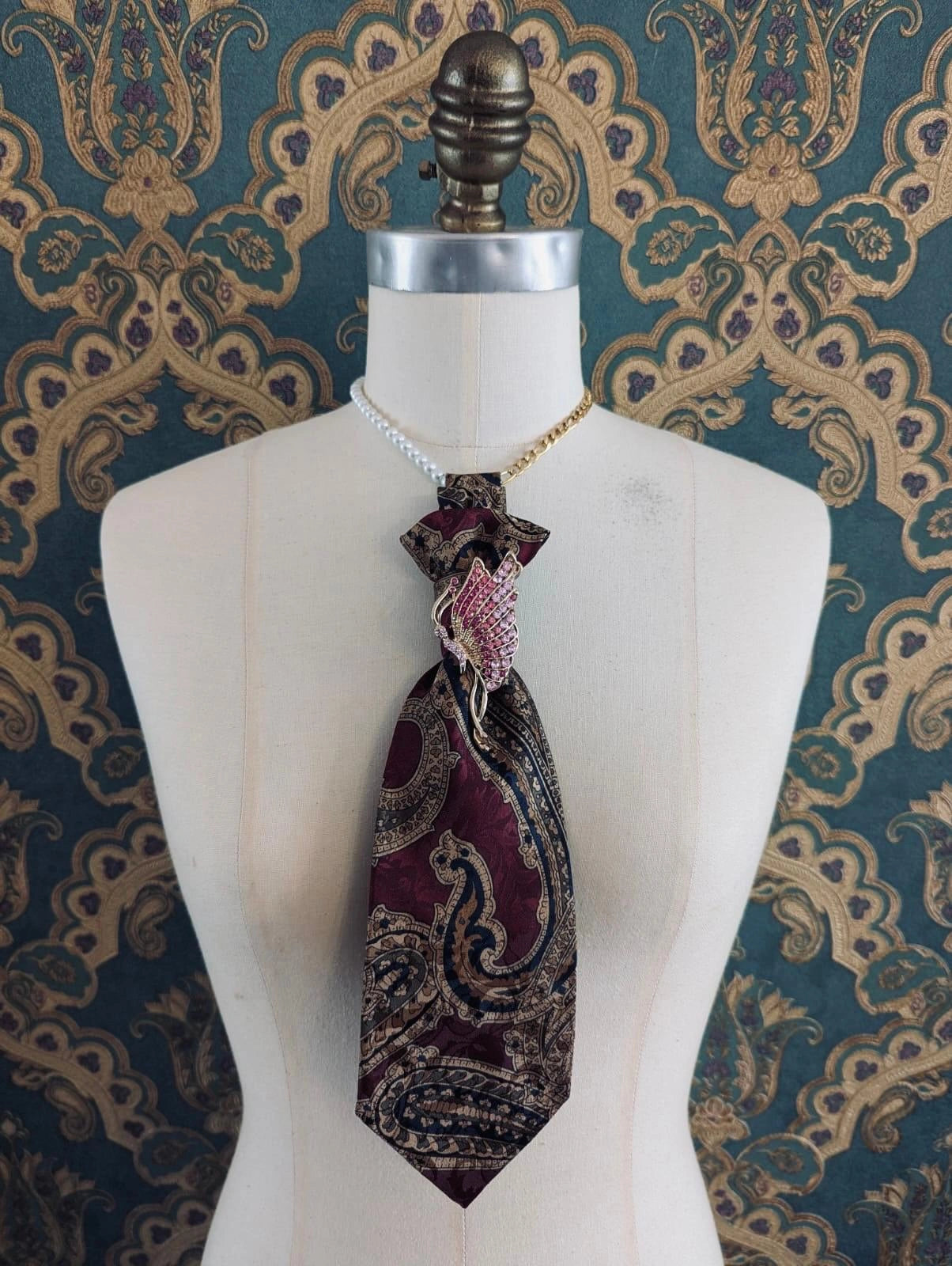 Broochella Montecristo cravatta vintage buurgundy paisley handmade up cycled tie brooch with vintage butterfly brooch embellishment. It is pinned to a mannequin The background is green and gold damask wallpaper. 