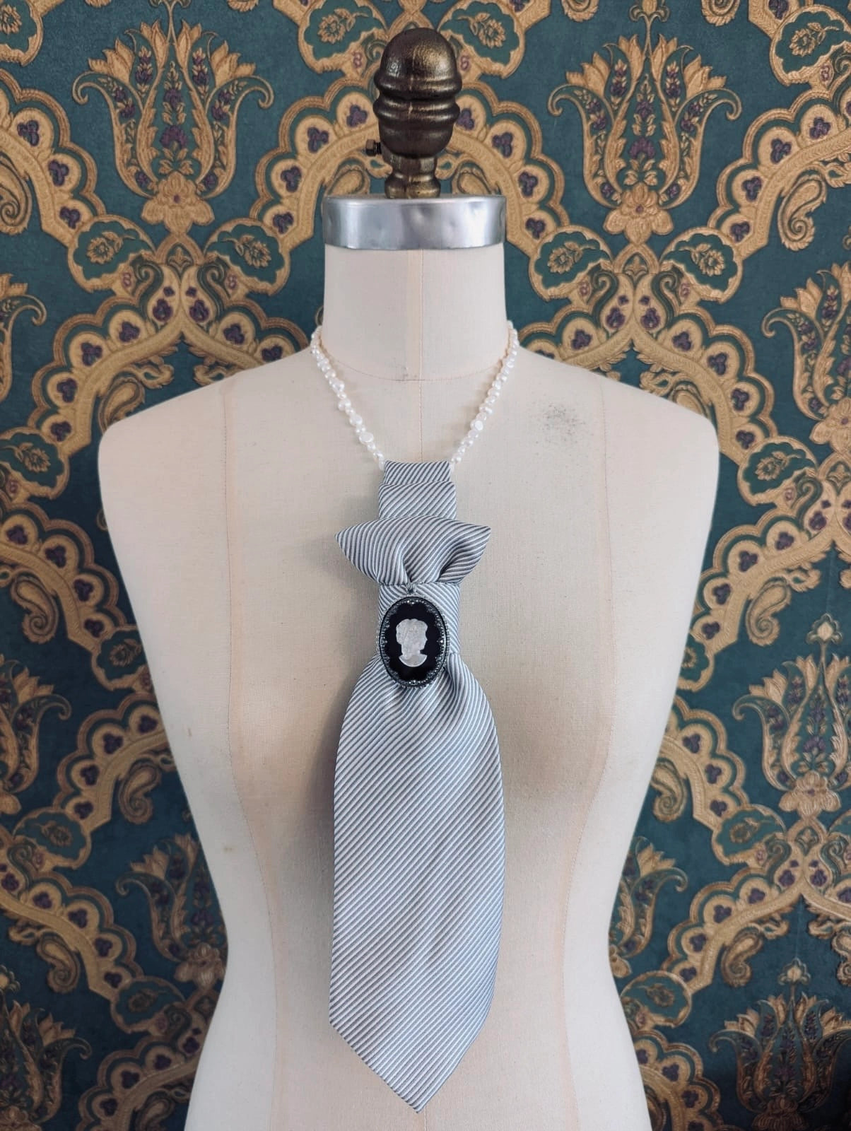 A Broochella Montecristo cravatta number 17 upcycled vintage tie brooch in grey and white stripe, with a vintage cameo embellishment. It is pinned on a cream-coloured dress form mannequin. The background is green and gold damask wallpaper. 