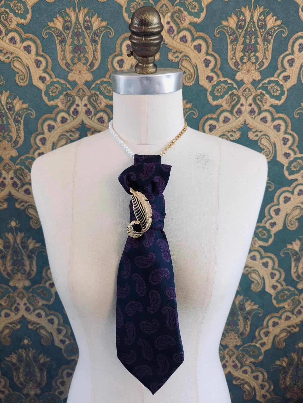 A Broochella Montecristo cravatta navy paisley upcycled, handmade tie brooch styled on a white dress form mannequin, featuring a gold leaf accent brooch and a gold chain, set against a teal and gold damask wallpaper background.