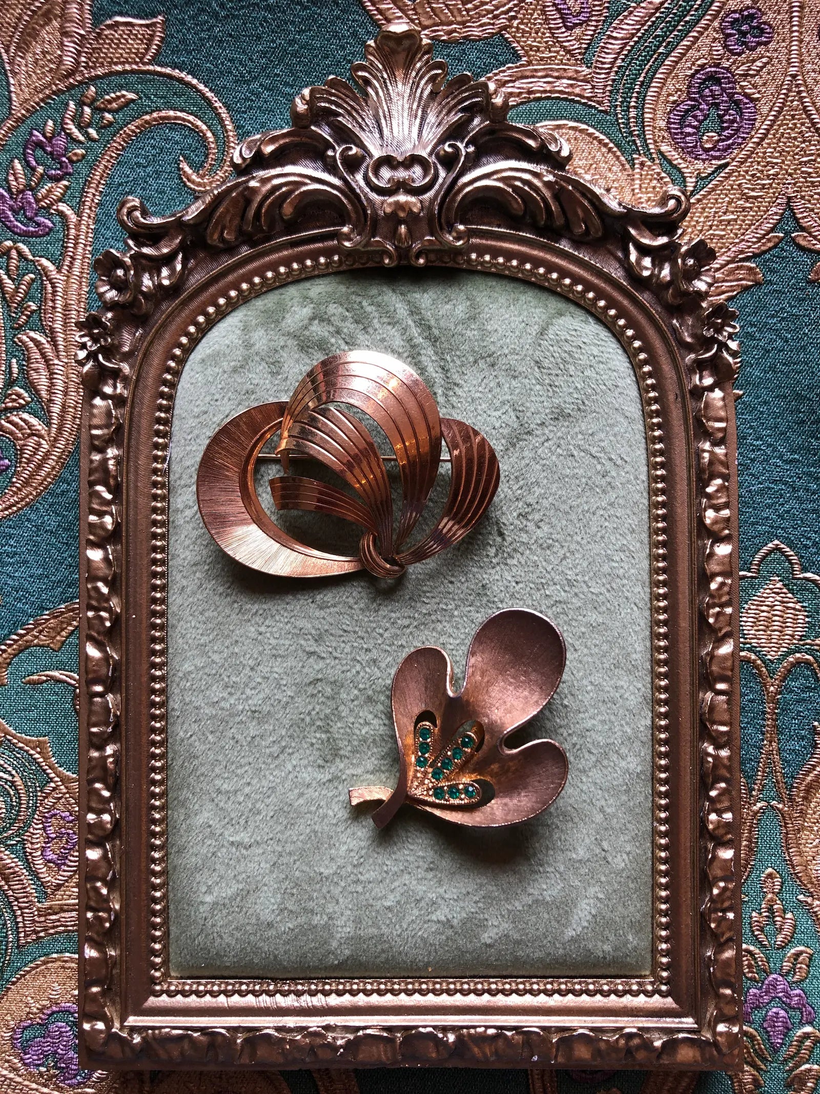 Broochella Movimento vintage modernist gold-tone brooch duo with an abstract ribbon brooch and a J.J. brooch featuring green crystals. Pictured inside an ornate gold frame, set against a patterned teal and gold fabric background.