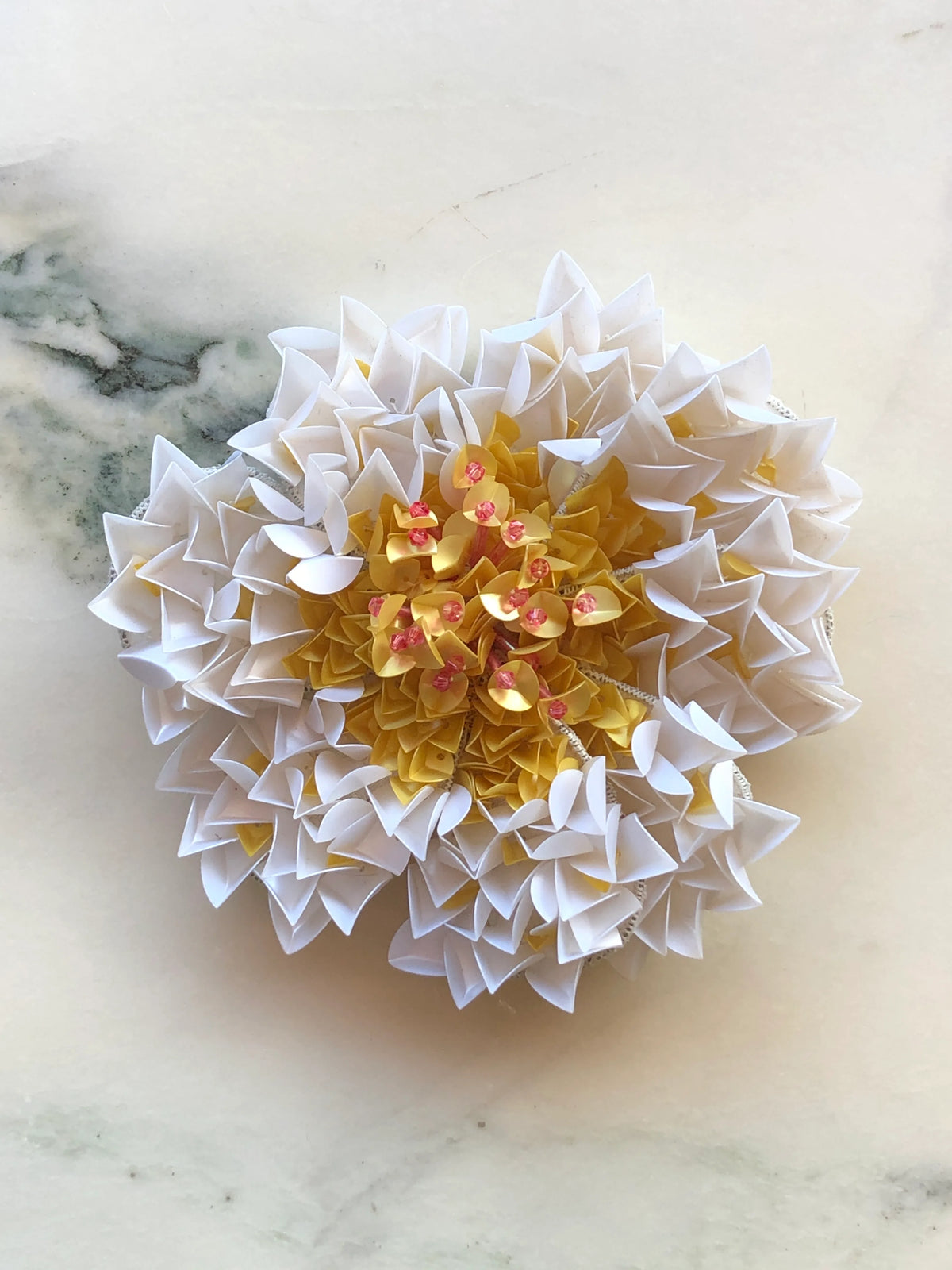 Broochella Narciso Flower Brooch featuring sculptural white petals with a golden yellow sequinned centre and pink bead details, photographed on a white marble background with soft grey veining.