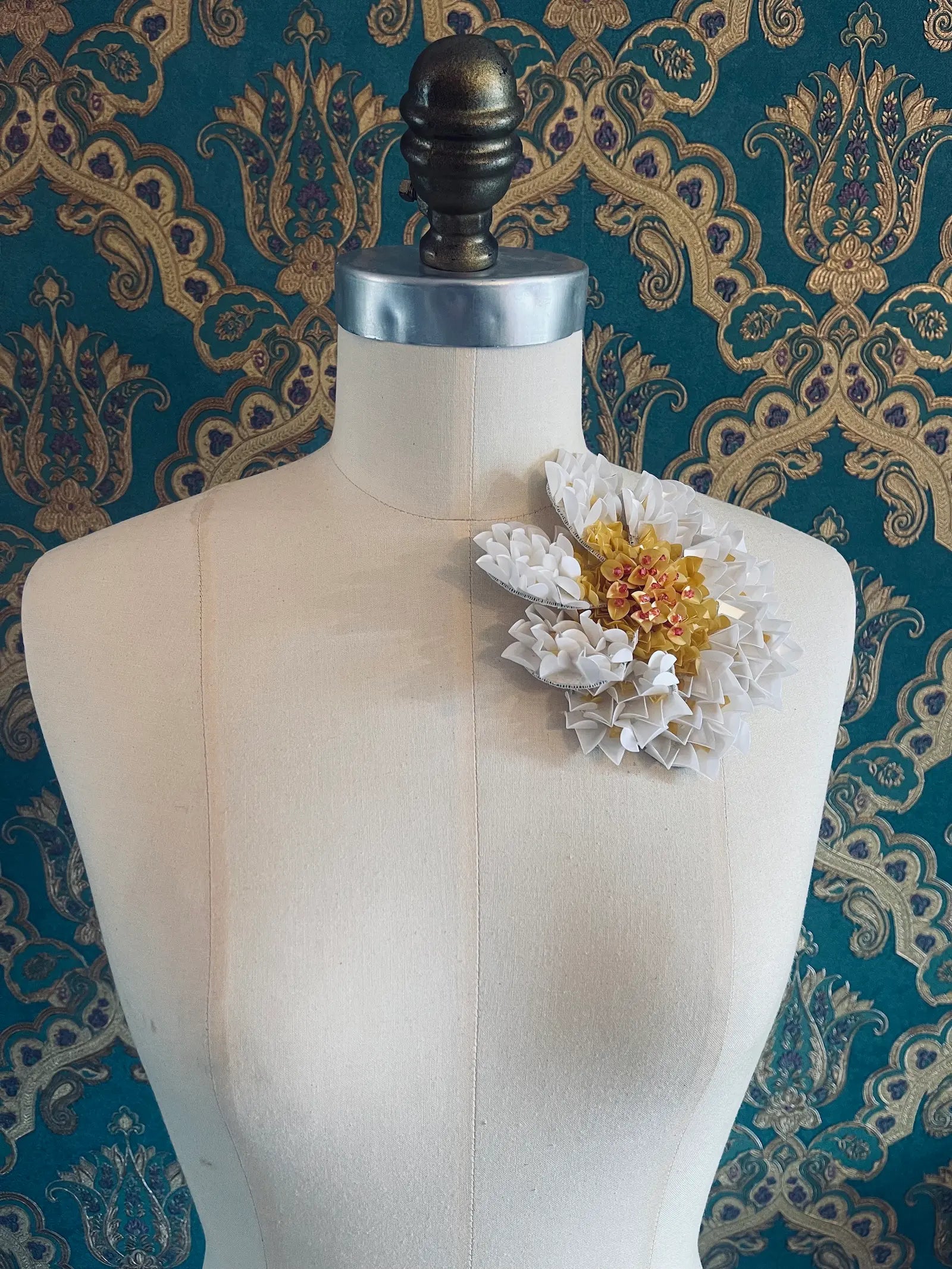 Broochella Narciso hand beaded sequin white flower brooch with a yellow centre. Pictured on a mannequin. The background is green and gold damask wallpaper. 