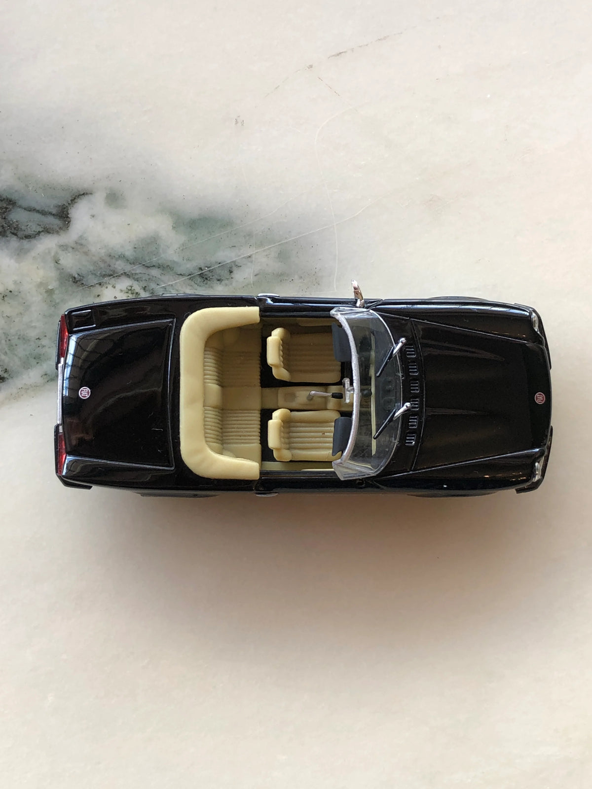 Broochella Nero Riviera No.84 Vintage Convertible Car Brooch photographed from above, showcasing its glossy black body, open-top design, cream ribbed seats, detailed interior, clear windscreen and black rear deck.