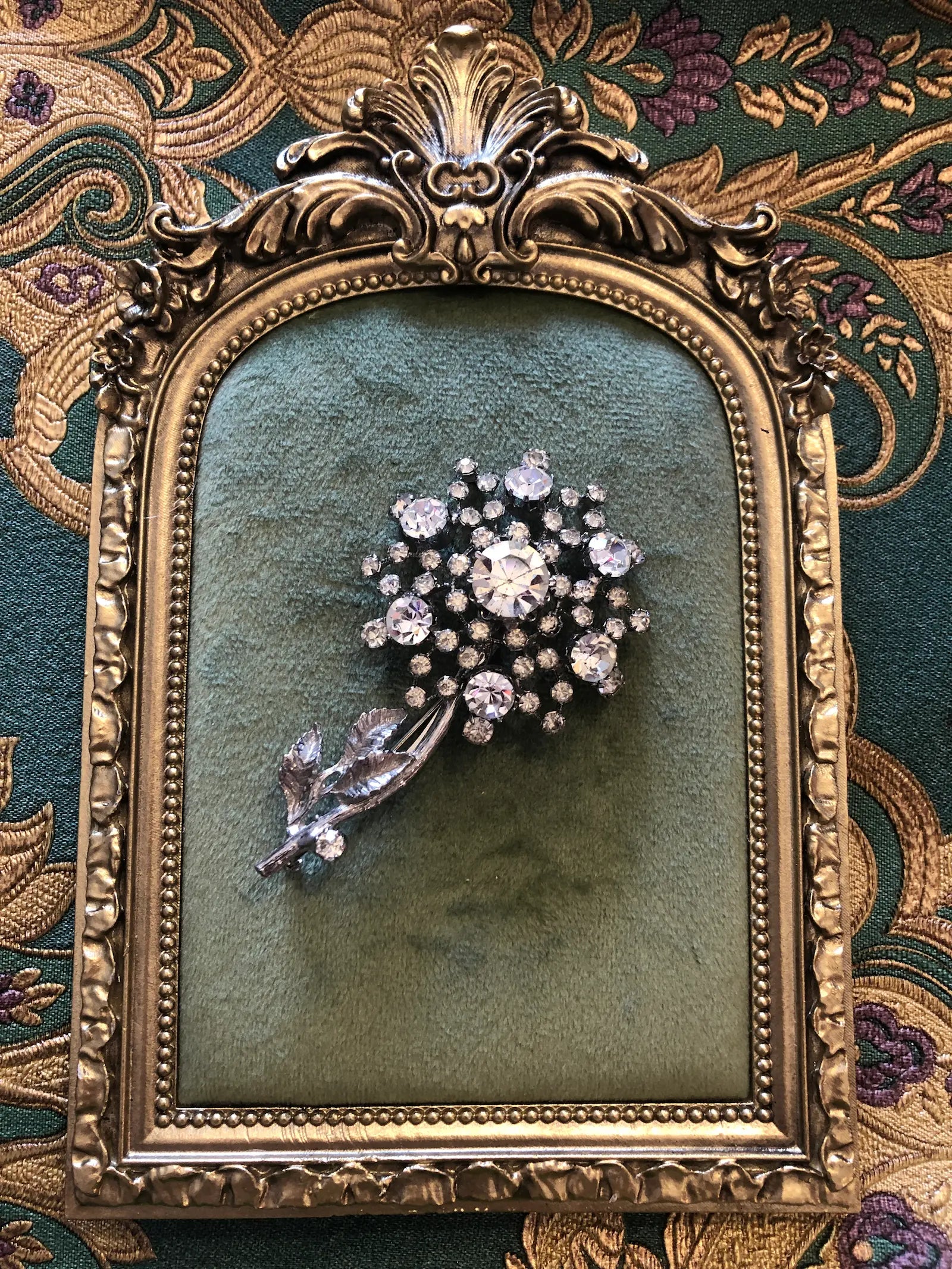 A Broochella Neve vintage flower brooch featuring a cluster of clear rhinestones and a leaf motif, displayed on a green velvet backing inside an ornate gold frame, set against a patterned green and gold fabric background.