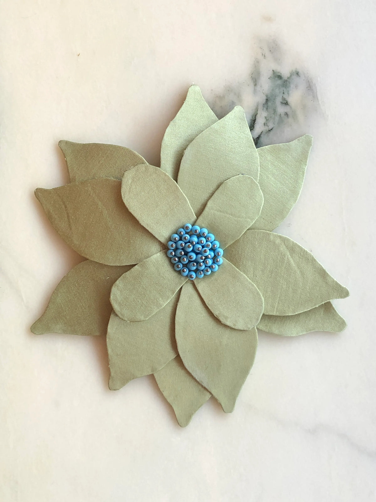 Broochella Ninfea Bejewelled flower brooch pin with layered pale sage green fabric petals and a turquoise blue beaded centre, photographed on a white marble background with soft grey-green veining.