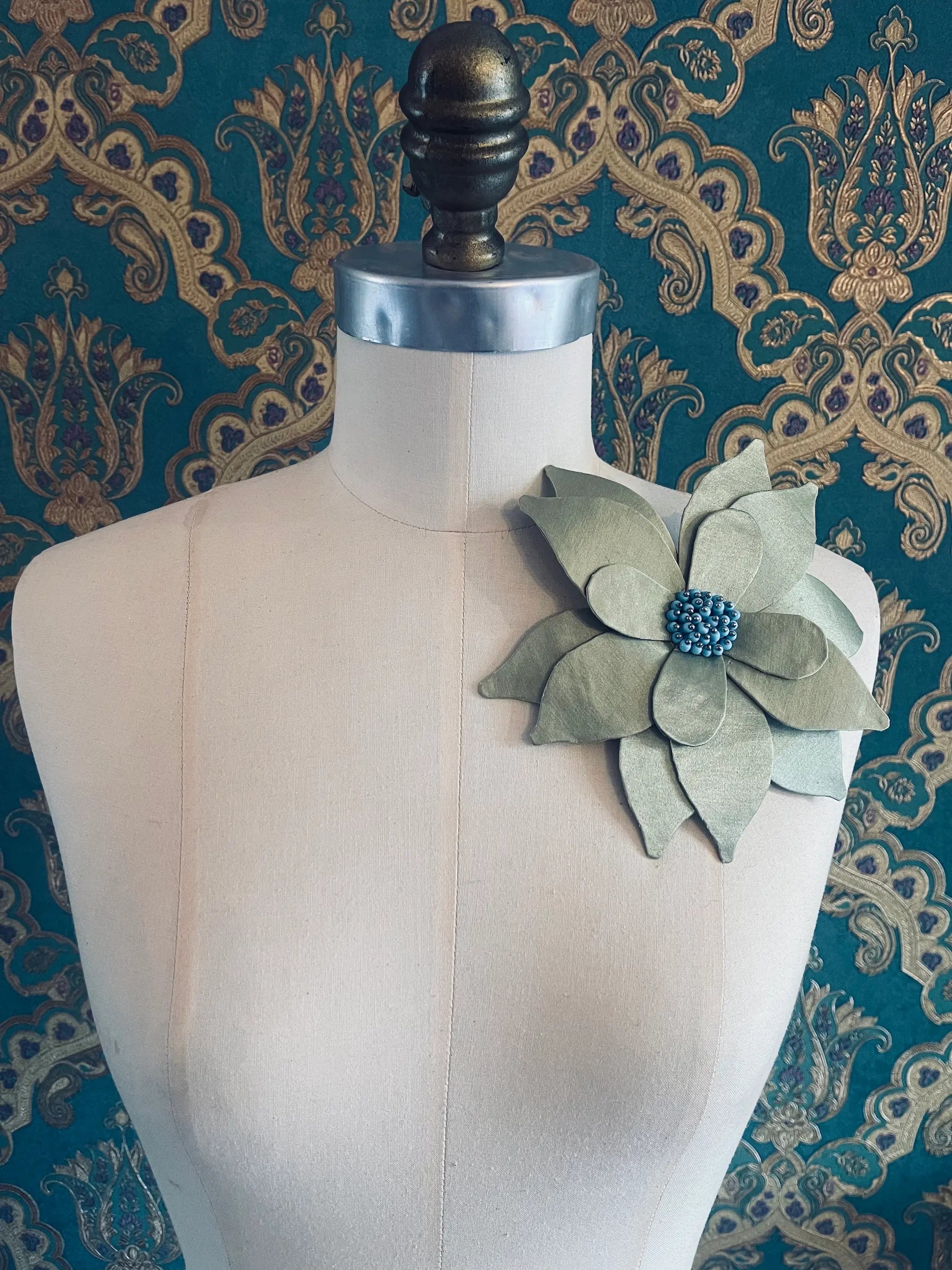 Broochella Ninfea handmade green fabric flower brooch with blue beaded centre. Pictured on a mannequin. The background is green and gold damask wallpaper