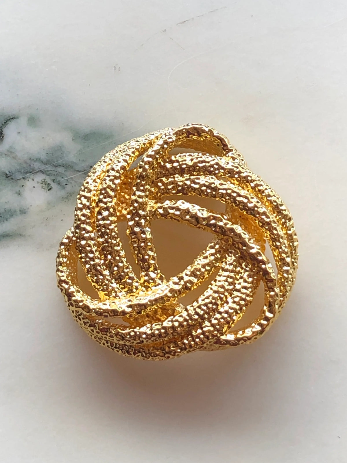 Broochella Nodo Golden Knot Brooch Pin featuring intertwined textured gold-tone loops in a sculptural knot design, photographed on a white marble background with soft grey-green veining.