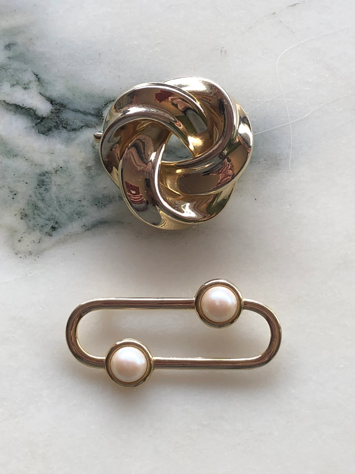 Broochella Noeud and Perle Vintage Brooch Duo featuring a sculptural polished gold-tone knot brooch and an elongated oval gold-tone brooch accented with two creamy faux pearls, photographed on a white marble background with soft grey-green veining.