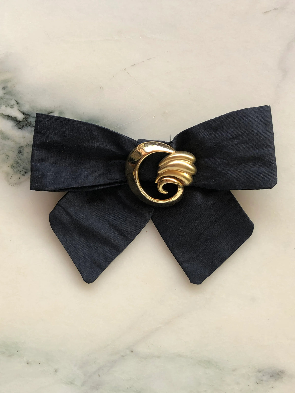 Broochella Noir Decadence Bow Brooch in black silk featuring a sculptural gold-tone vintage-style centre embellishment, photographed on a pale white marble background with grey veining.