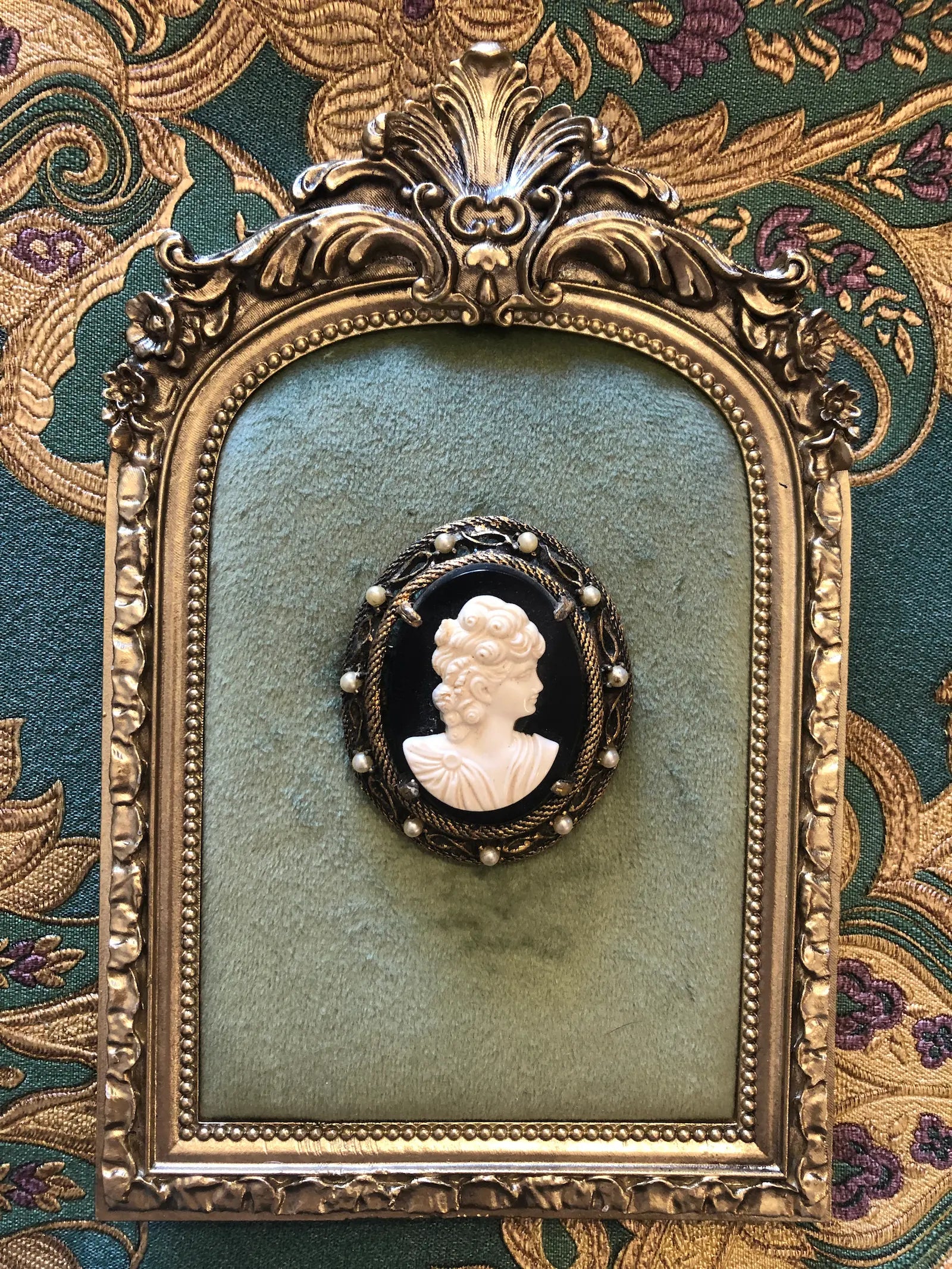 A 5.5cm x 4.5cm vintage cameo brooch featuring a carved white classical portrait on a deep black ground set within a gold-tone rope twist filigree frame with seed pearl accents. Photographed on a green velvet tray with a gold frame. The background is green and gold damask wallpaper. 