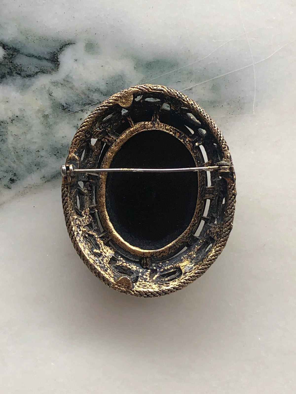 Broochella Noire Vintage Cameo Brooch Pin back view showing the oval black cameo setting, ornate antique gold-tone frame and pin fastening, photographed on white marble with soft grey-green veining.