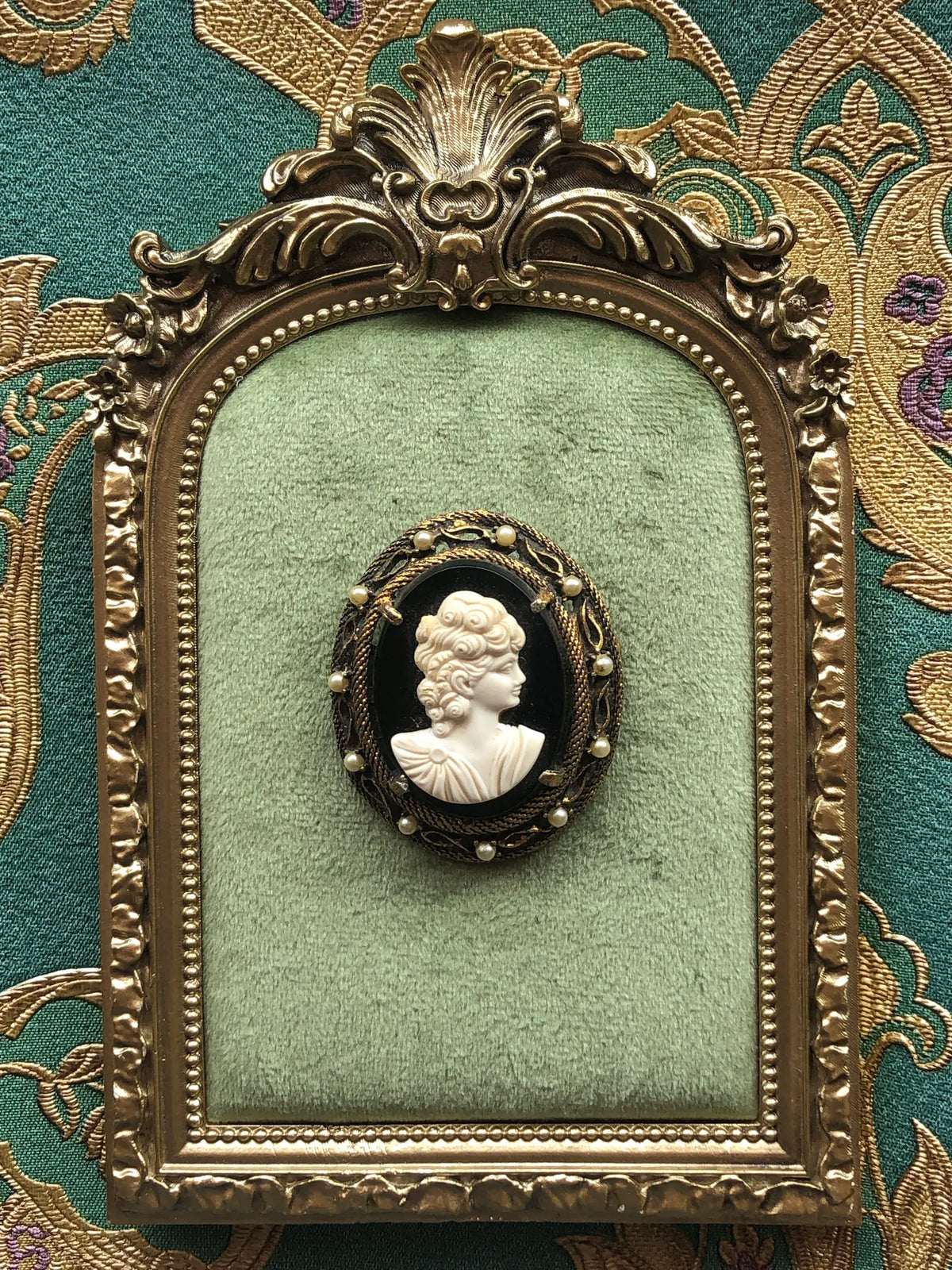 Broochella Noire Vintage Cameo Brooch Pin featuring a sculpted ivory female profile on a black oval background, surrounded by an ornate antique gold-tone frame with pearl accents, displayed on sage green velvet in an antique gold frame against green and gold patterned fabric.