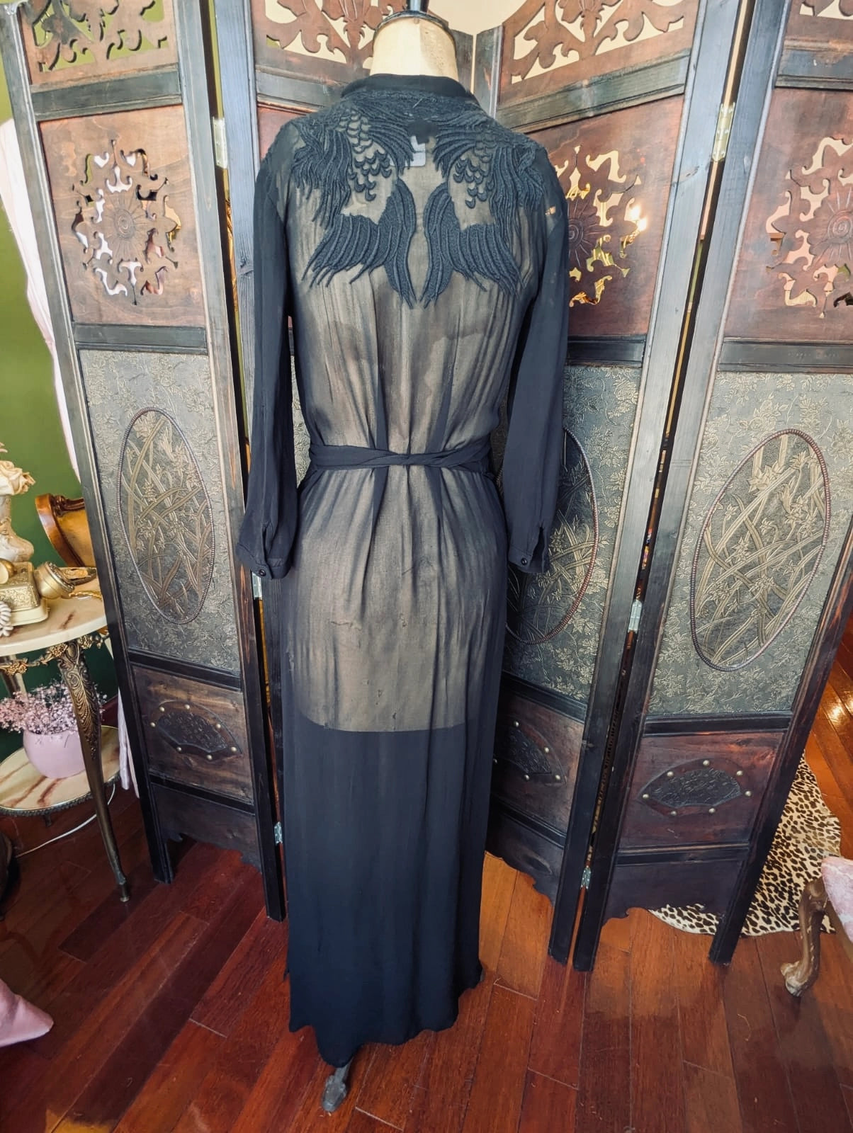 The back view of the Broochella Nü Denmark size small Long sheer black dress with embroidered lace detailing on a mannequin against a decorative screen.