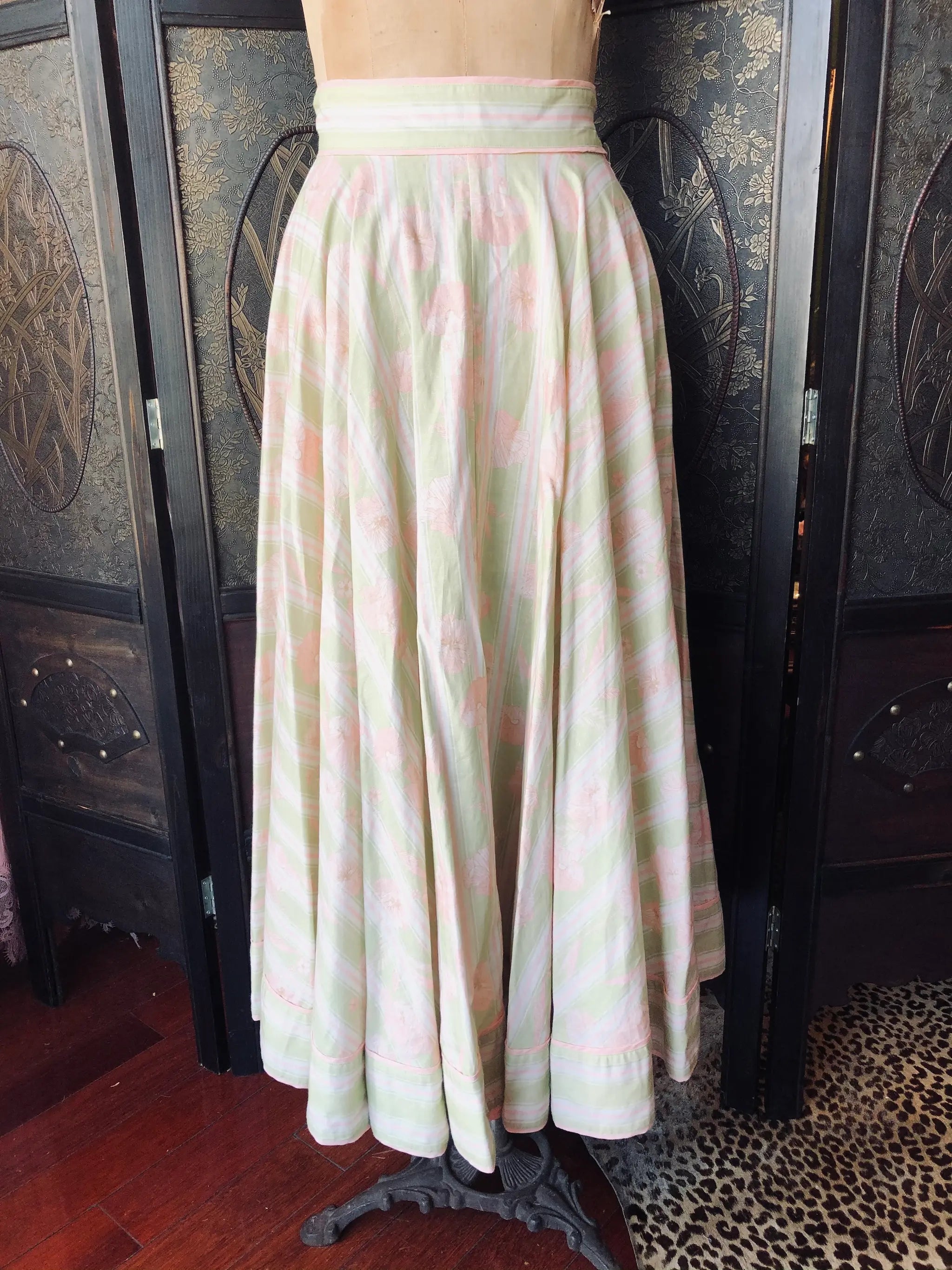 A Broochella Odetr high-waisted, full-length A-line skirt with a pink and green floral and striped pattern on a cream base, displayed on a dress form mannequin in front of a dark wooden folding screen.