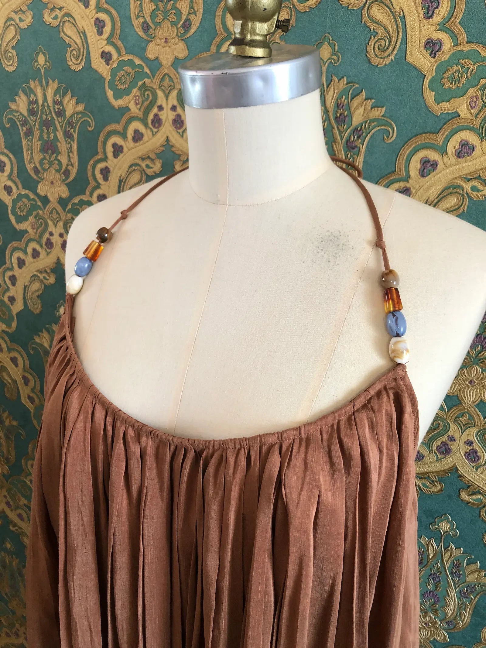 Odet Tropicale cocoa linen and silk sunray-pleated sundress with beaded halter straps, size Small. Pictured on a mannequin. The background is green and gold damask wallpaper.