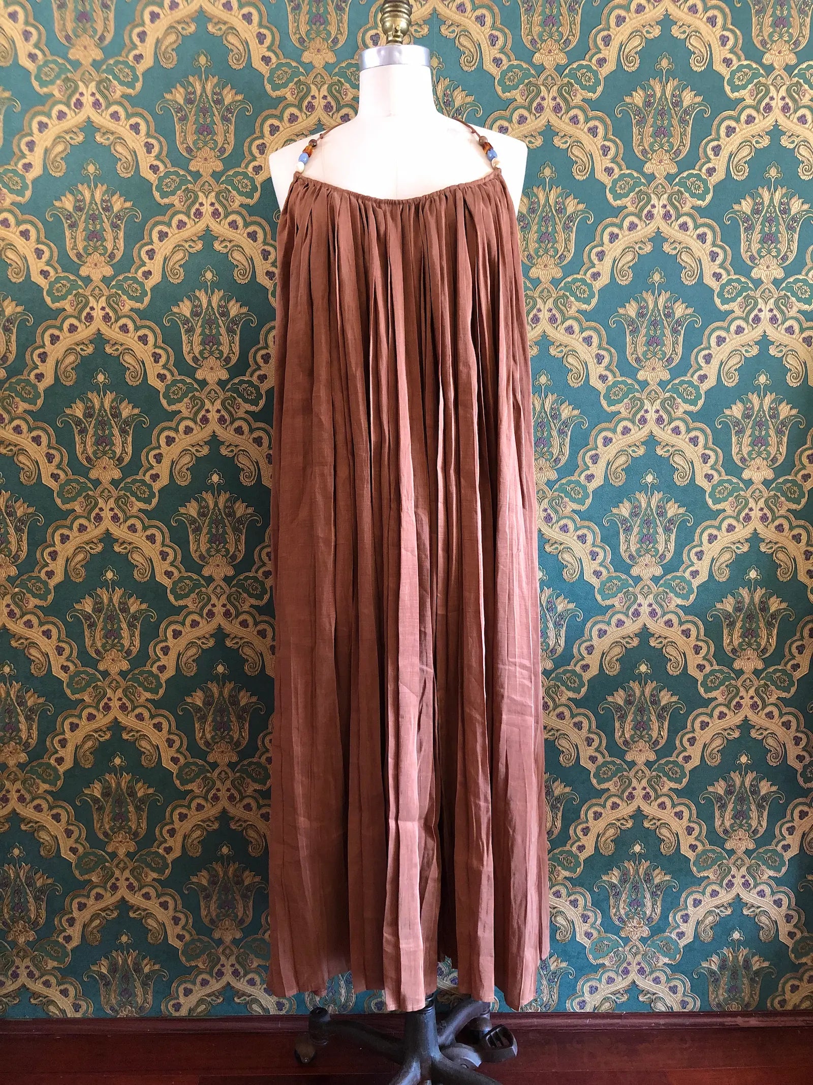Odet Tropicale cocoa linen and silk sunray-pleated sundress with beaded halter straps, size Small. Pictured on a mannequin. The background is green and gold damask wallpaper.