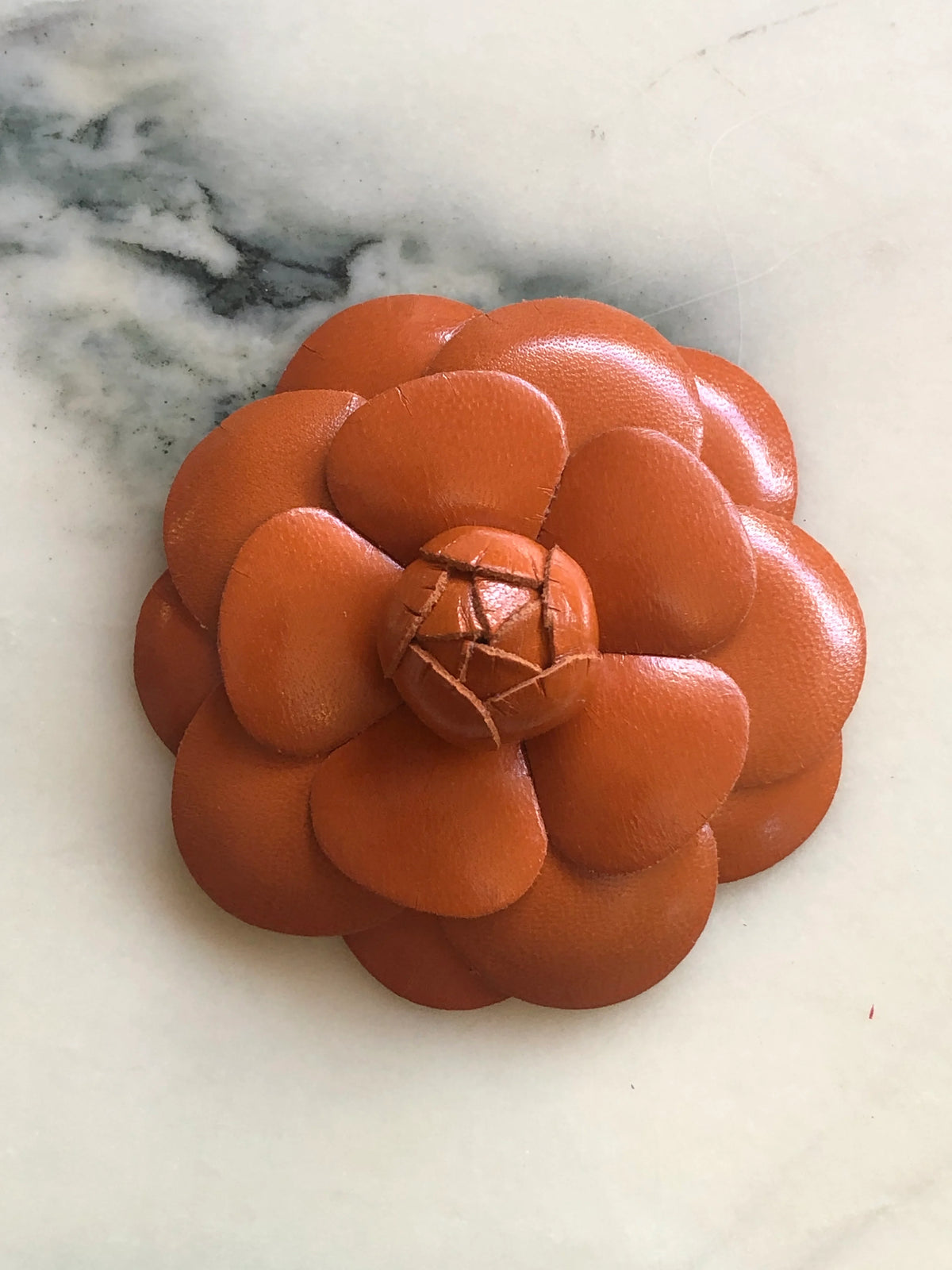 A Broochella Camelia handmade orange leather flower-shaped accessory brooch,  resting on a white marble surface.