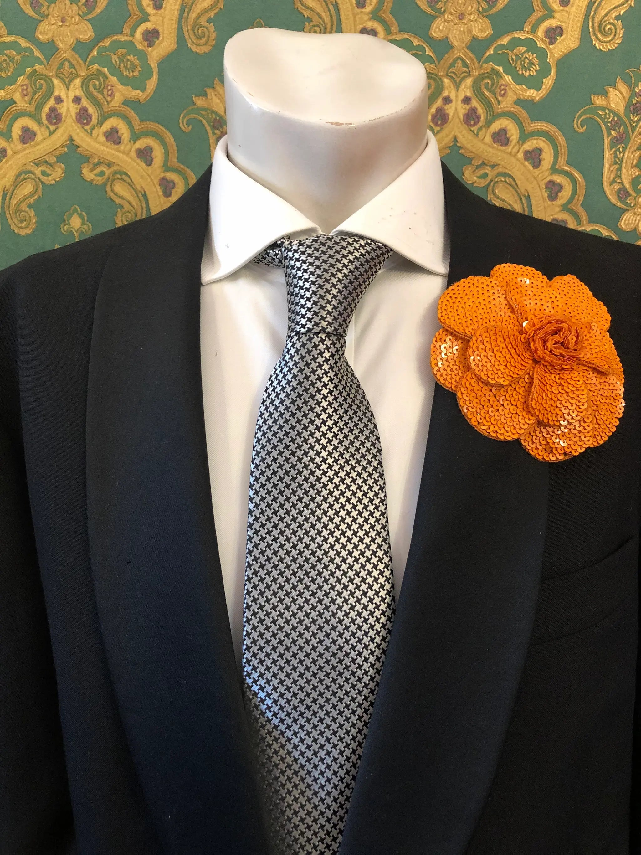 A men's formal accessory set displayed on a white mannequin torso, featuring a black suit jacket, a white dress shirt, a silver and black checkered tie, and an orange Men's Broochella Scintillante sequinned flower brooch lapel pin against a green and gold patterned wallpaper background.