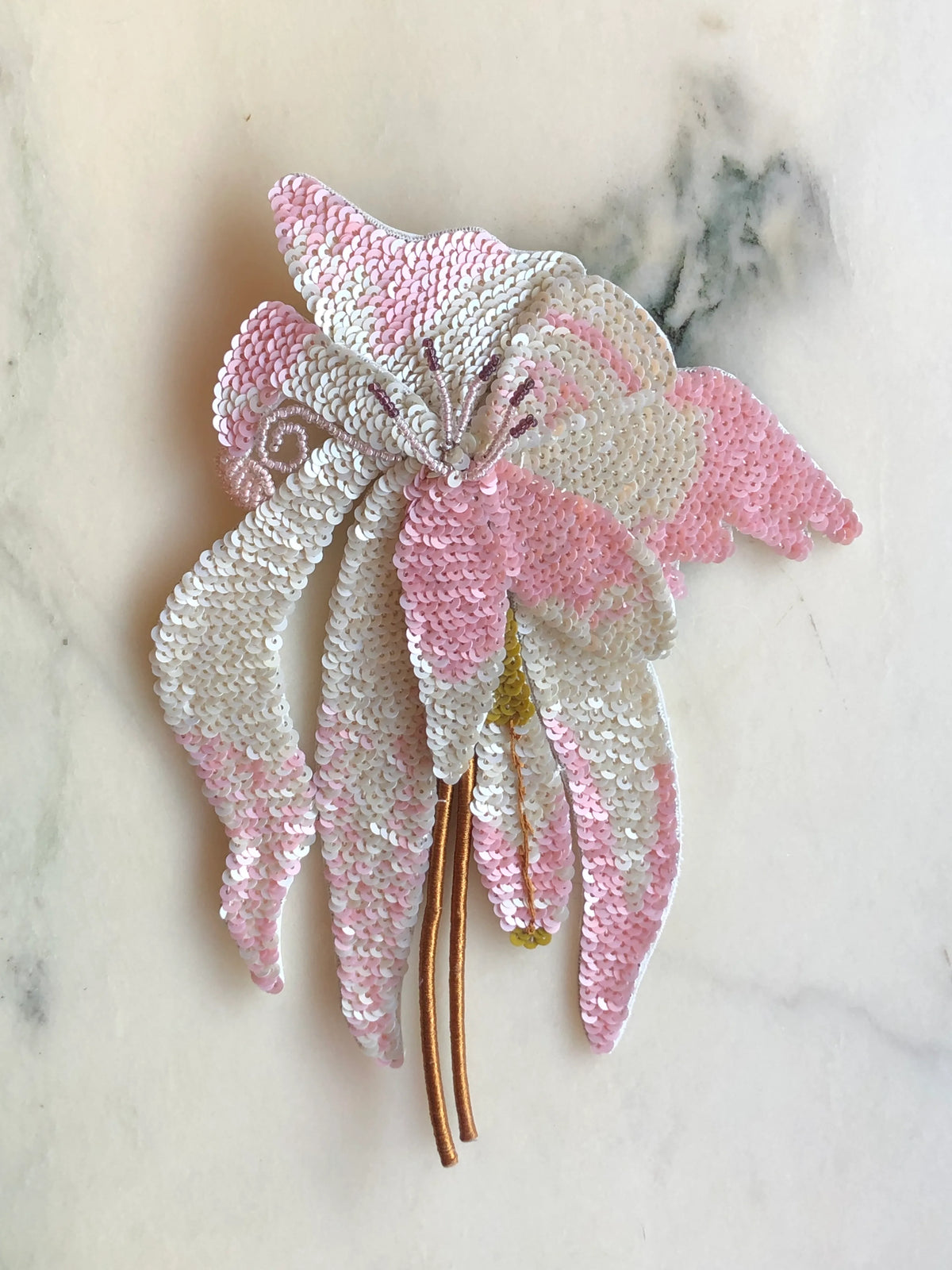 Broochella Orchid Sequin Flower Brooch Pin embellished with soft pink and white sequins, featuring sculptural orchid petals, delicate beaded details and long gold stems, photographed on a white marble background with soft grey-green veining.